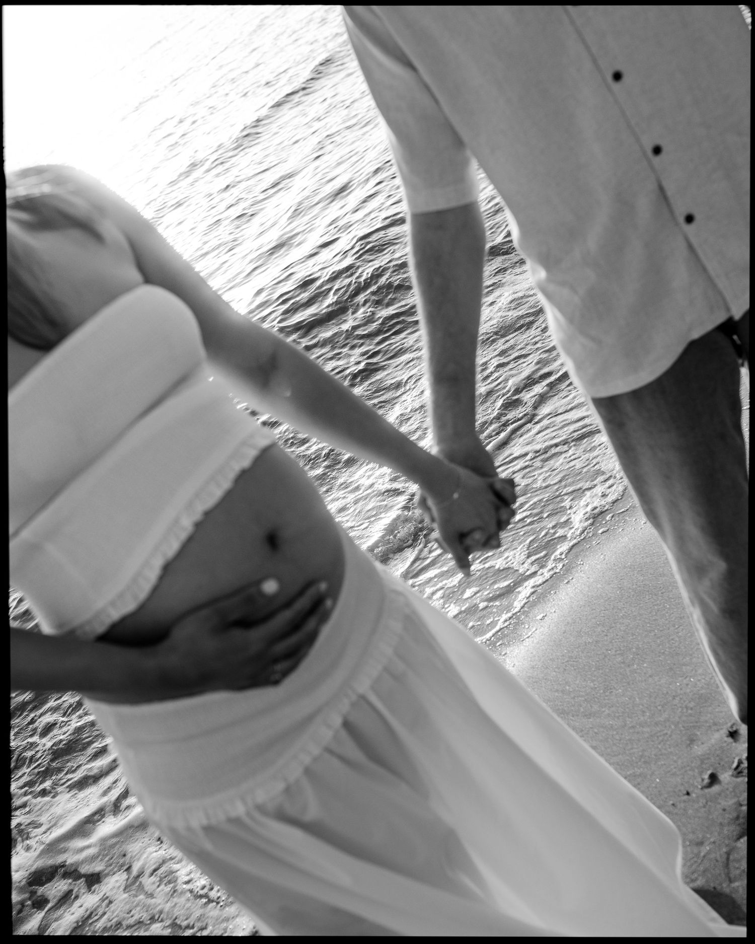 Black and white photo of pregnant couple holding hands at beach shoreline.
