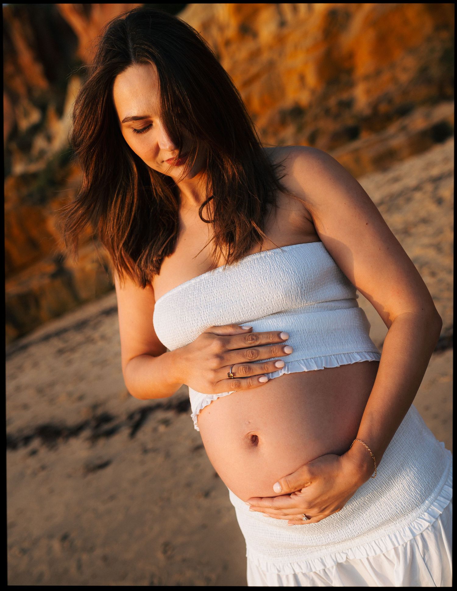 Close-up of pregnant woman in white bandeau top gently holding her baby bump on sandy beach.