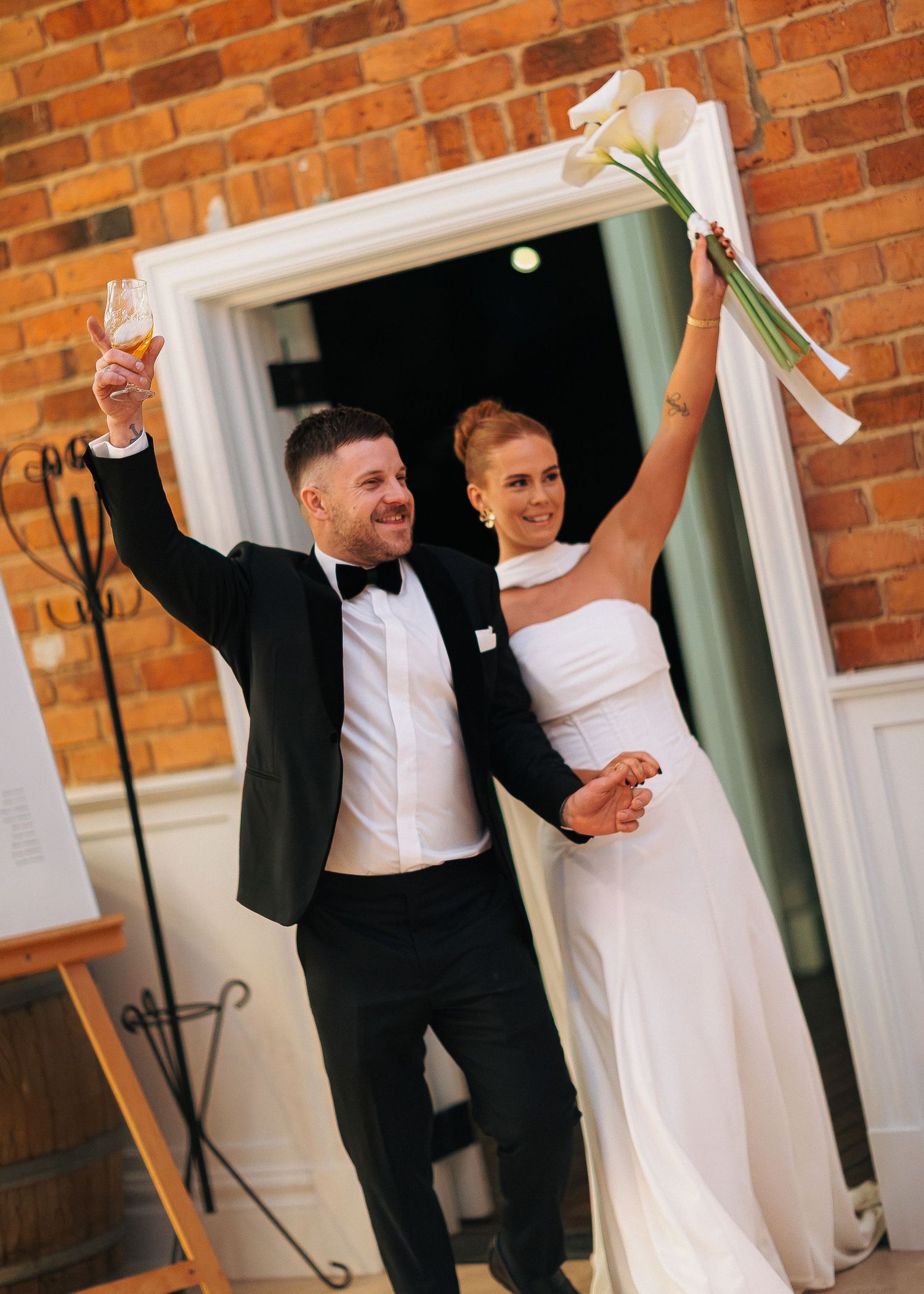 Joyful newlyweds celebrating their wedding, groom in tuxedo raising champagne, bride in white gown holding calla lilies bouquet.