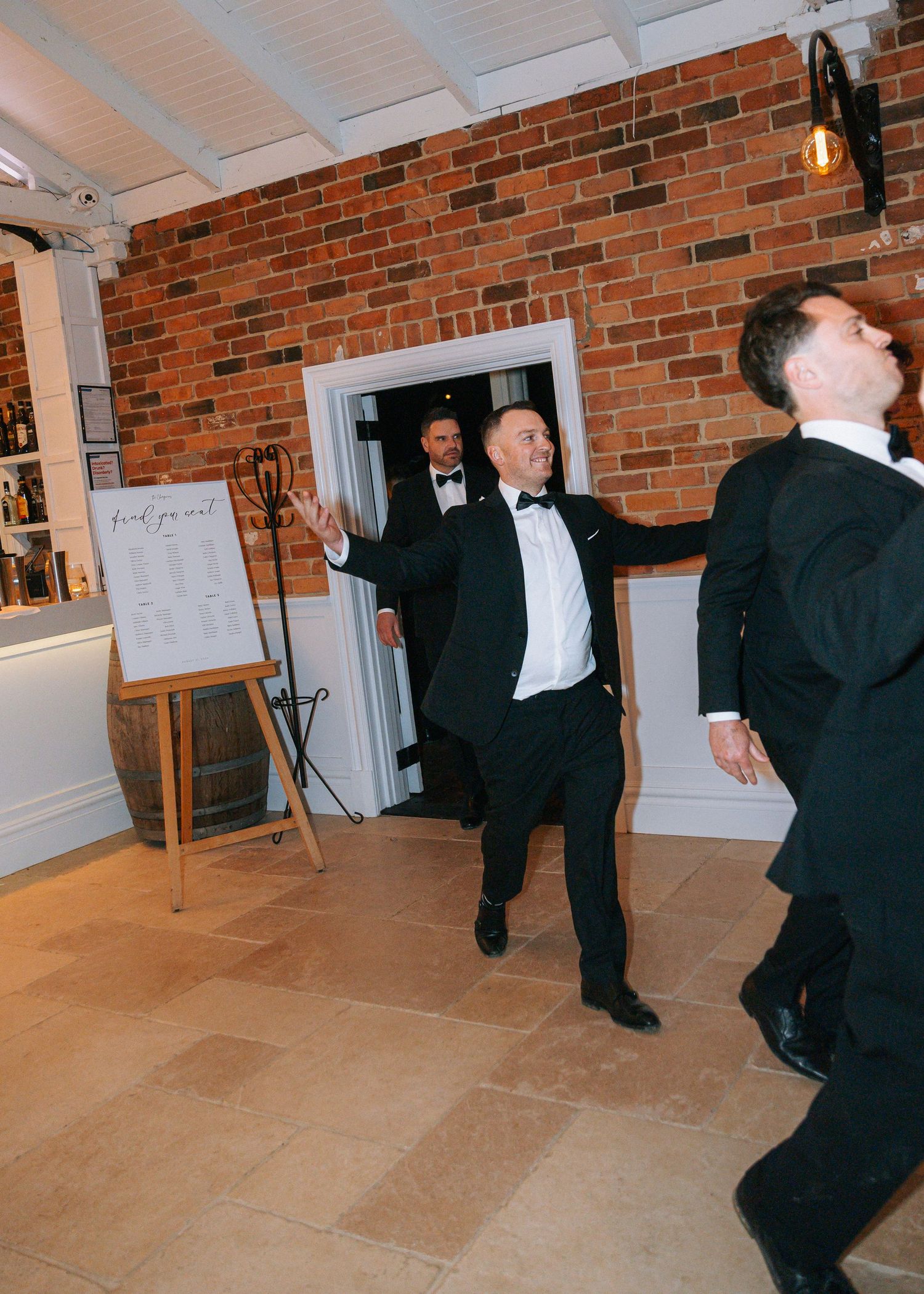 Groomsmen in black tuxedos making grand entrance at wedding reception venue with brick walls.