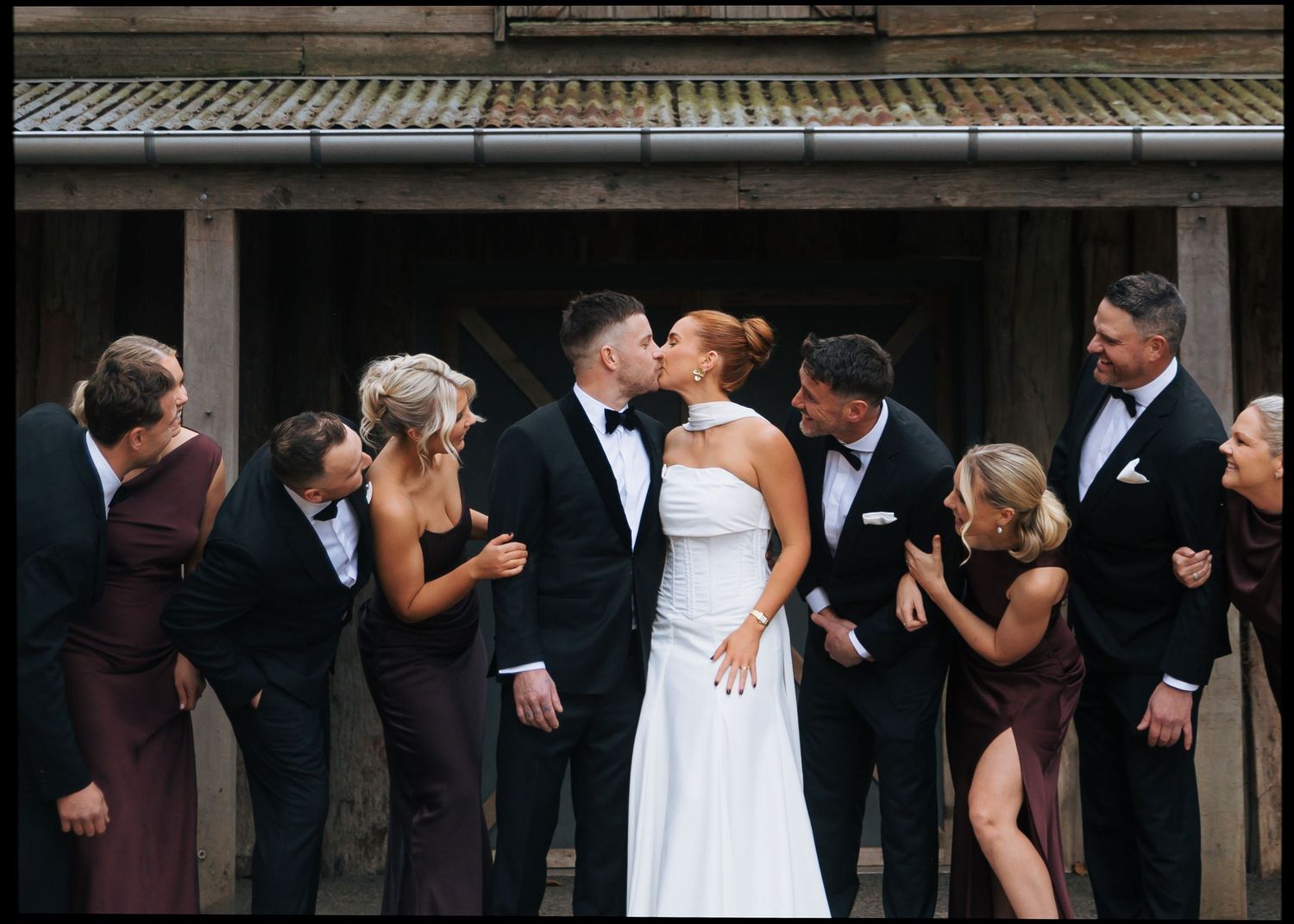 Wedding couple kissing surrounded by bridal party in black tuxedos and burgundy dresses outside rustic venue.