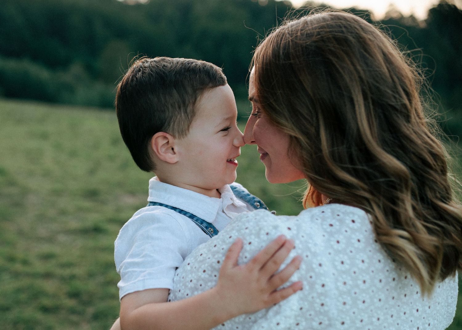 A smiling mother holds her laughing toddler boy nose-to-nose in a tender outdoor moment.