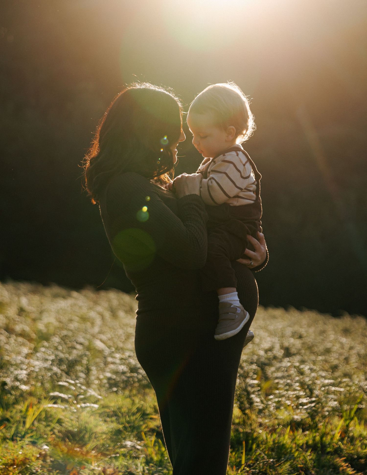 Silhouetted mother and toddler sharing a tender moment in radiant golden backlit field.