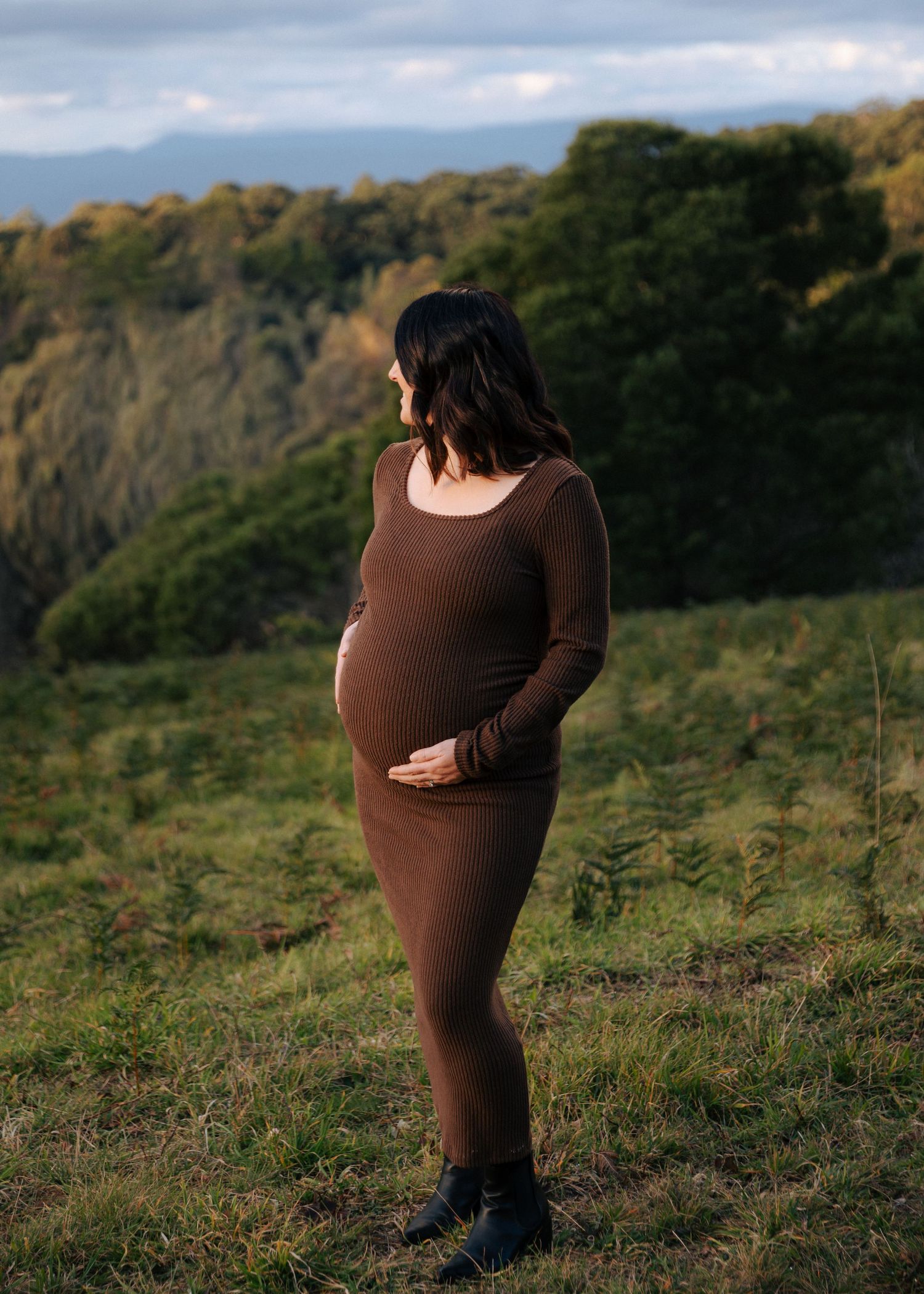 Pregnant woman in brown dress cradling baby bump in scenic outdoor setting.