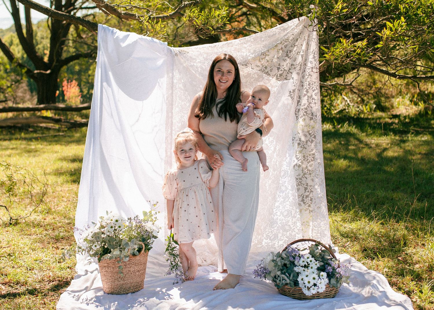 A mother stands with her two young children in front of a lace backdrop surrounded by flower baskets outdoors.