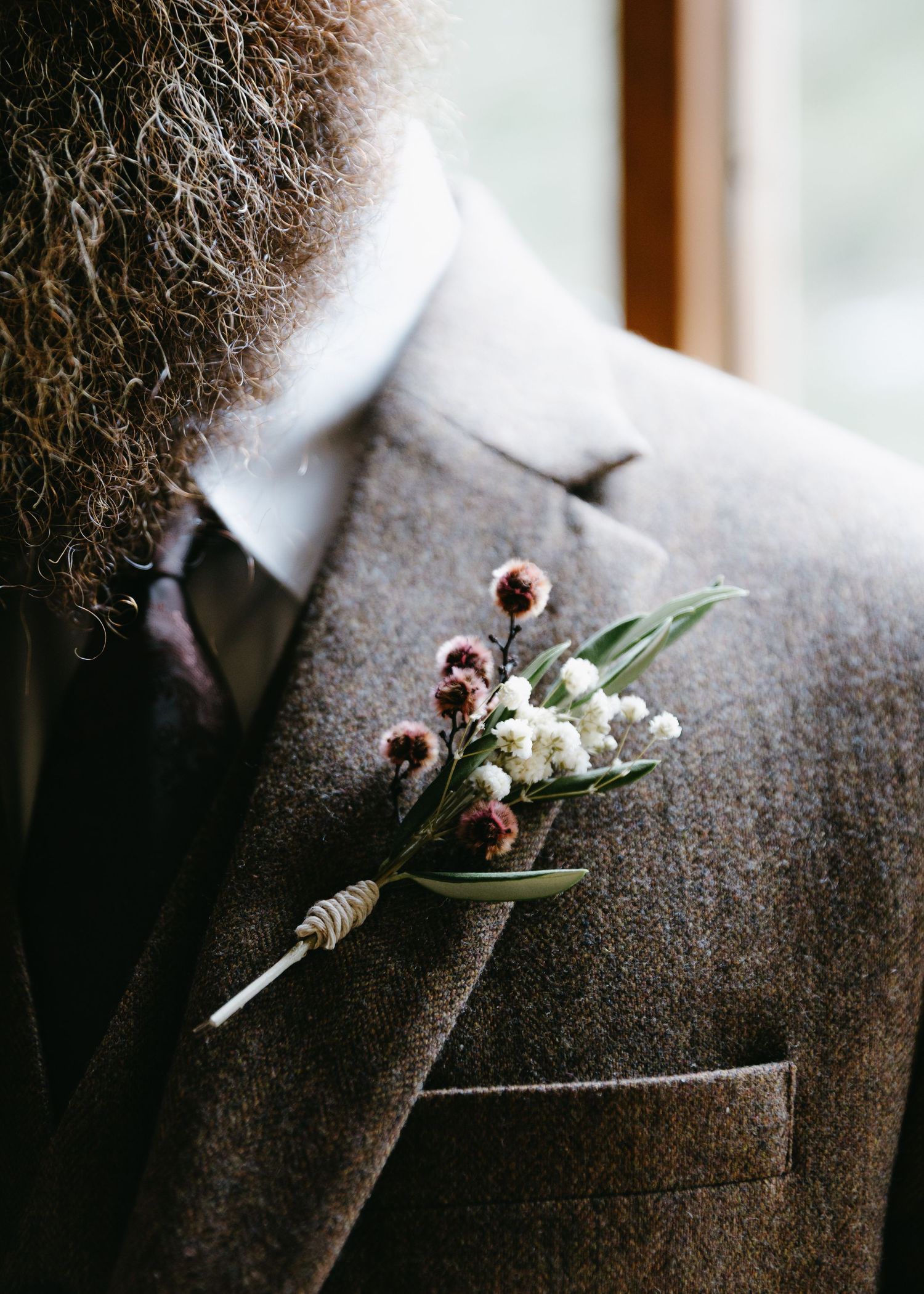 Close-up of a rustic boutonniere with white and purple wildflowers on a brown tweed wedding suit lapel.