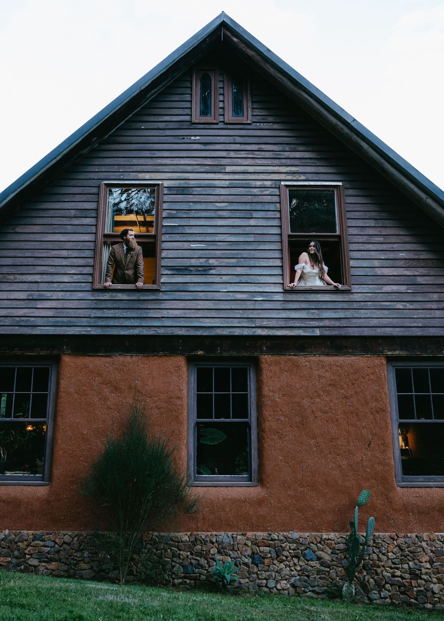 Bride and groom posing in separate windows of a rustic wooden barn venue at dusk.