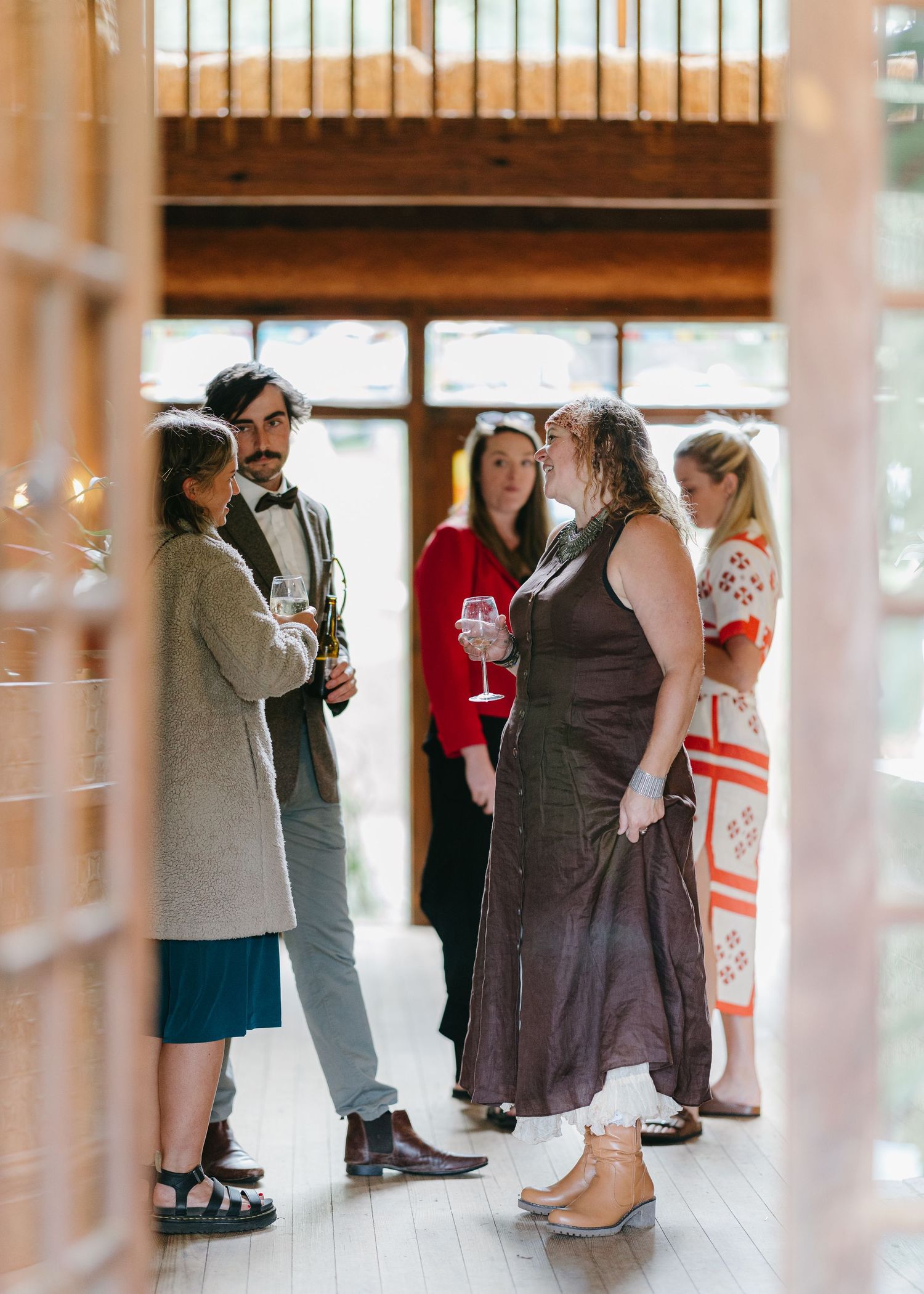 Elegantly dressed wedding guests socializing indoors at a rustic venue, holding wine glasses.