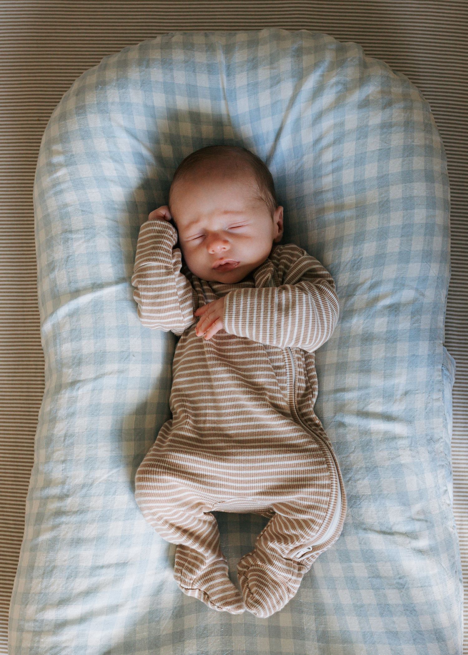 Newborn baby in striped onesie sleeping in a blue gingham baby lounger.