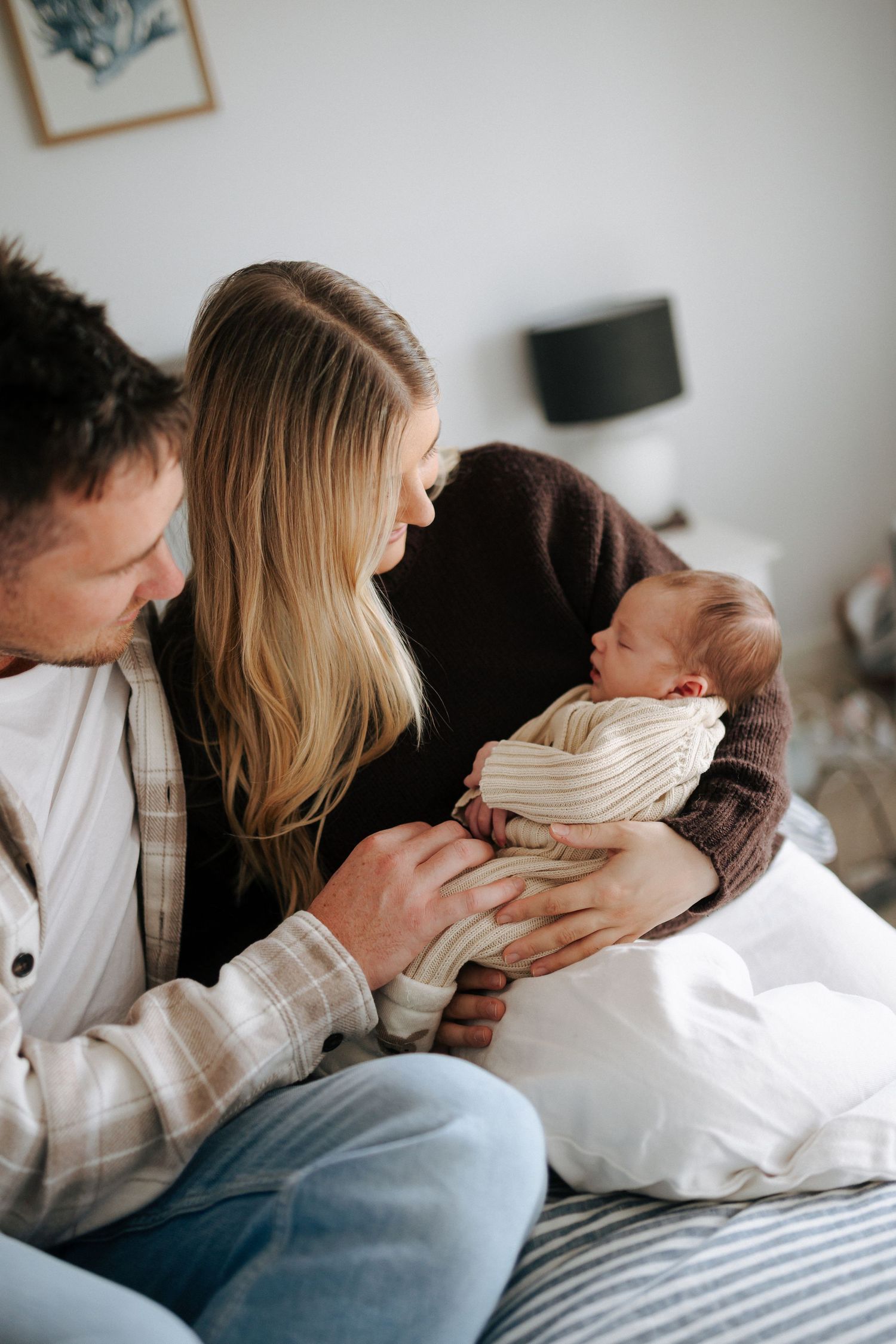 New parents sitting on bed together admiring their sleeping newborn baby wrapped in cream knit outfit.