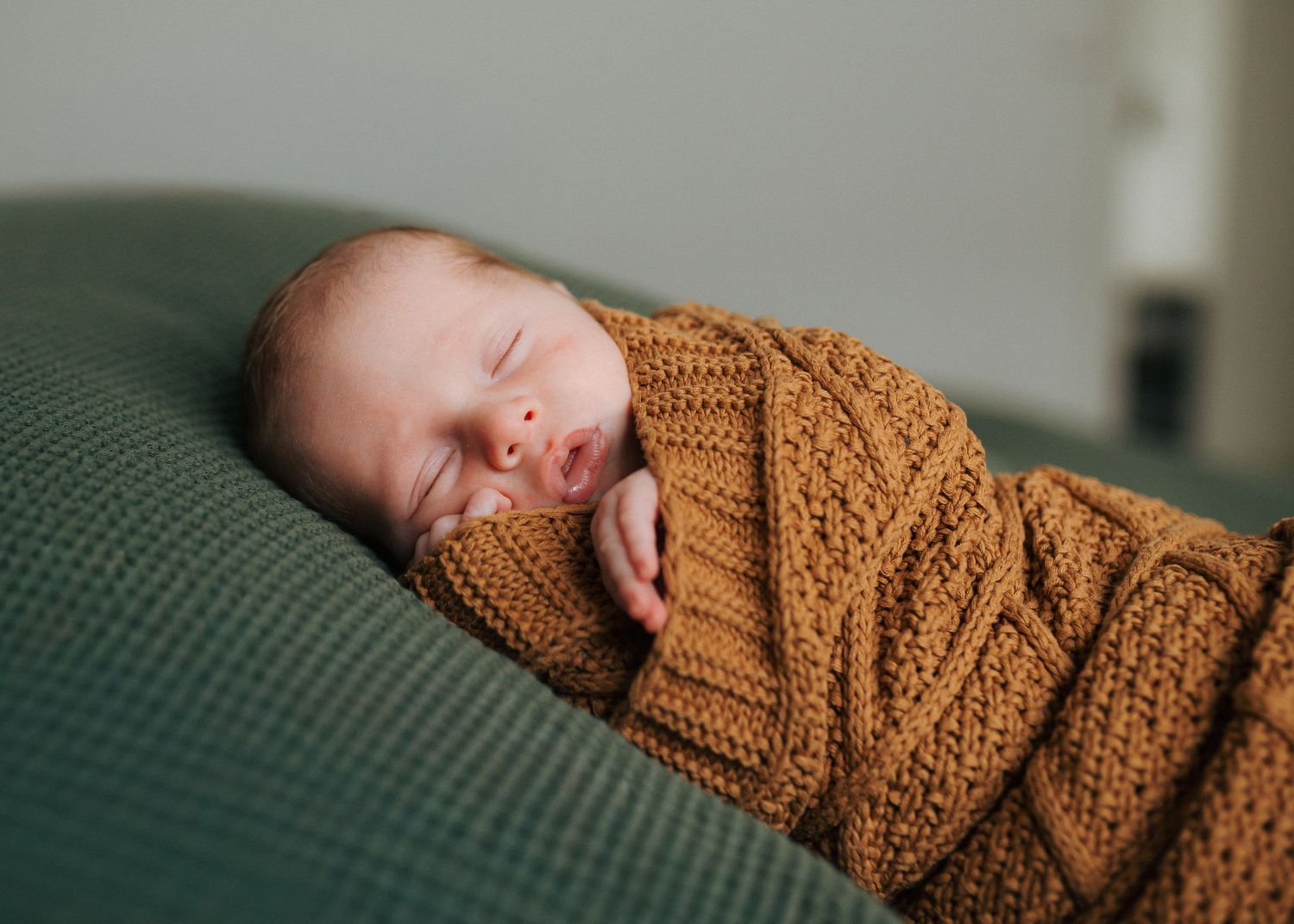 Sleeping newborn baby swaddled in a warm brown knit blanket on a green cushion.
