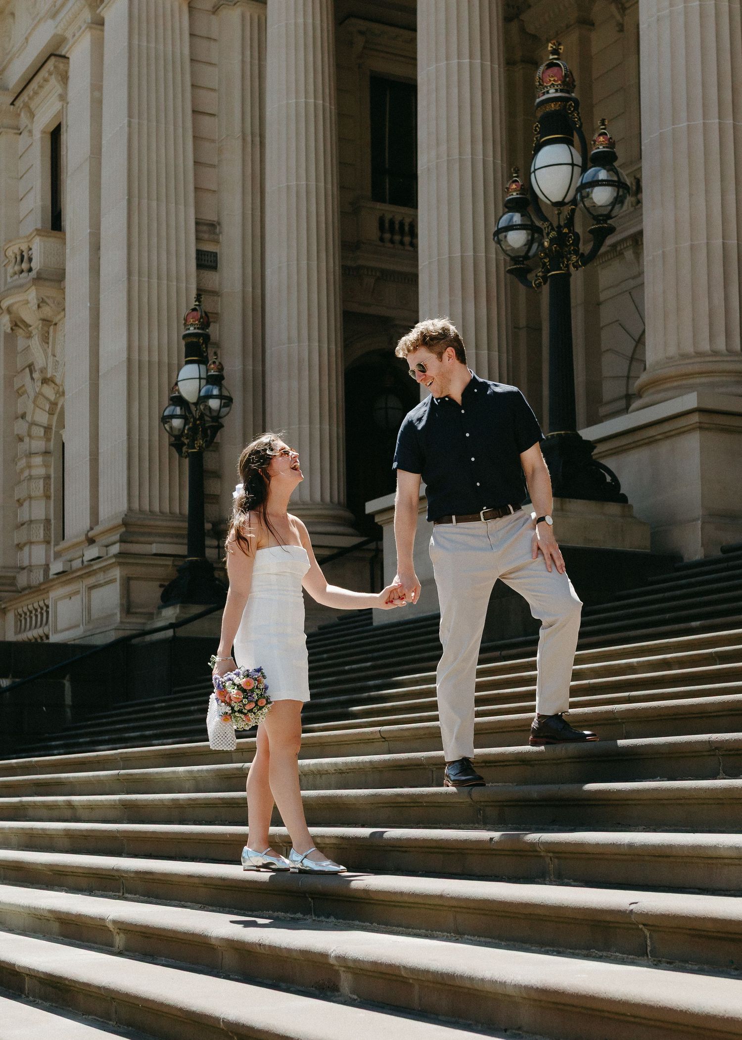 Couple holds hands walking down grand stone steps of historic building on sunny day.