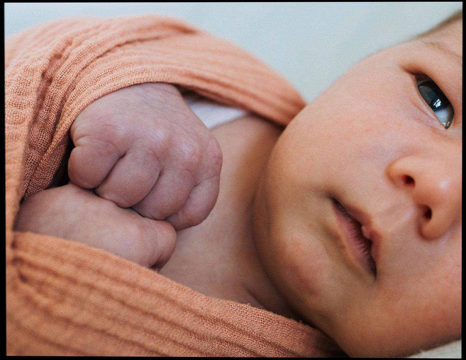 Close-up of a newborn baby wrapped in a soft peach knit blanket, with a tiny clenched fist raised near the face and eyes gazing sideways.
