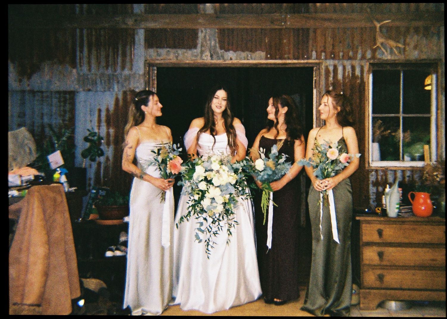 Bride and three bridesmaids in mismatched green and white gowns holding bouquets in rustic barn.