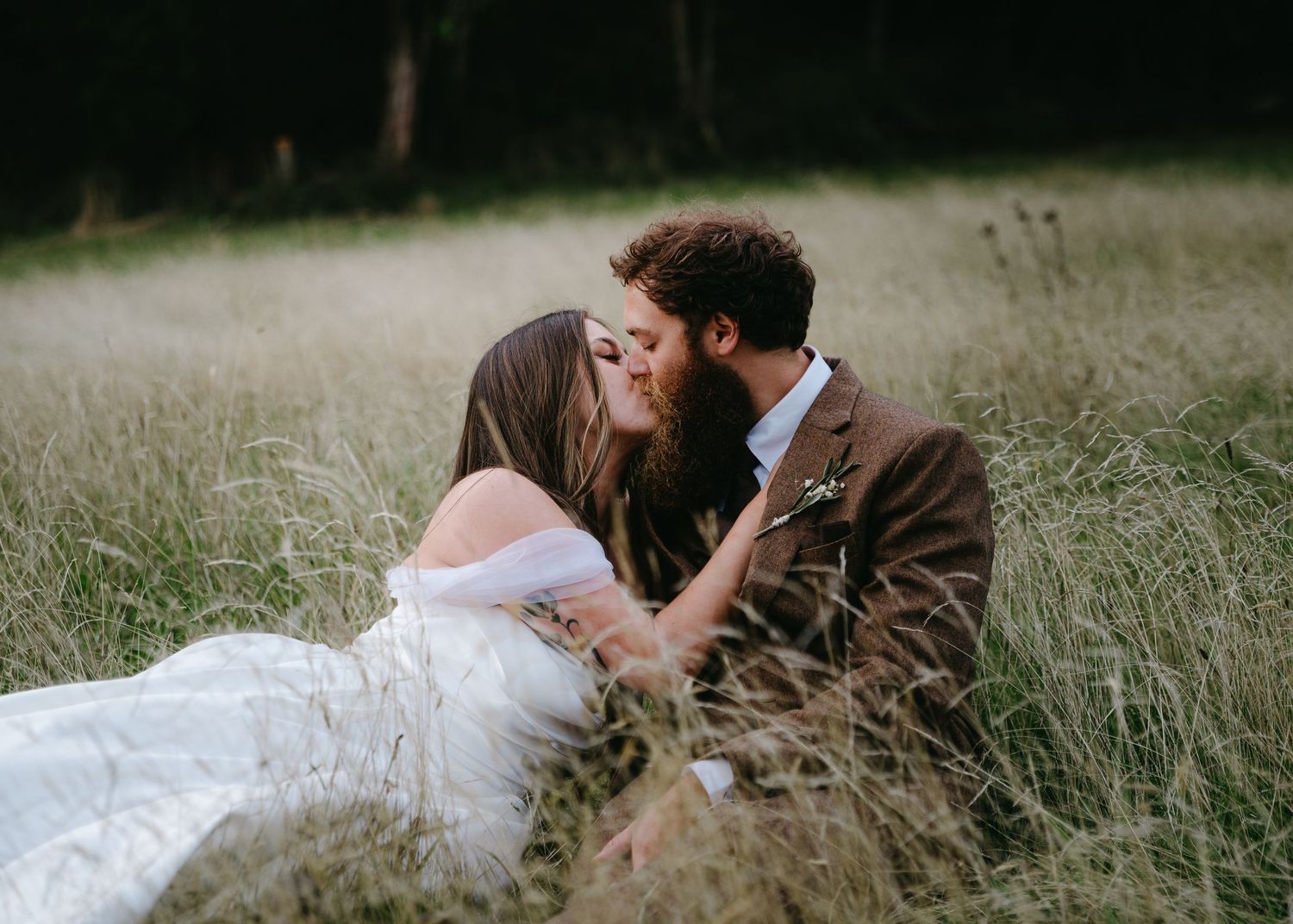 Bride and groom sharing a kiss while sitting in tall golden grass during outdoor wedding photoshoot.