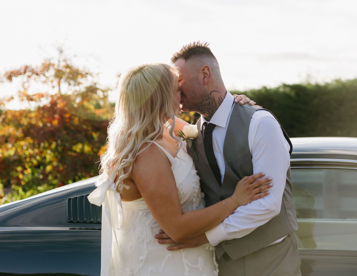 Newlywed couple sharing a romantic kiss outdoors, bride in lace gown and groom in gray vest, golden hour lighting.jpg