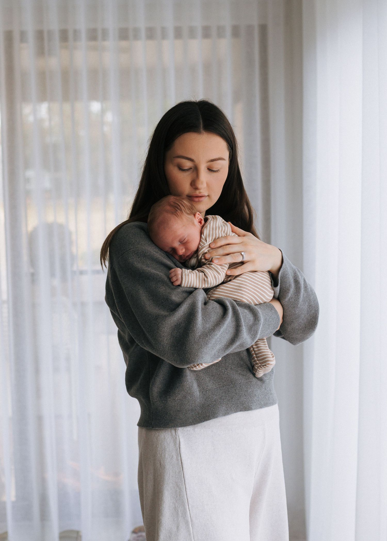 A dark-haired mother in a grey sweater holds her sleeping newborn close to her chest by a window.