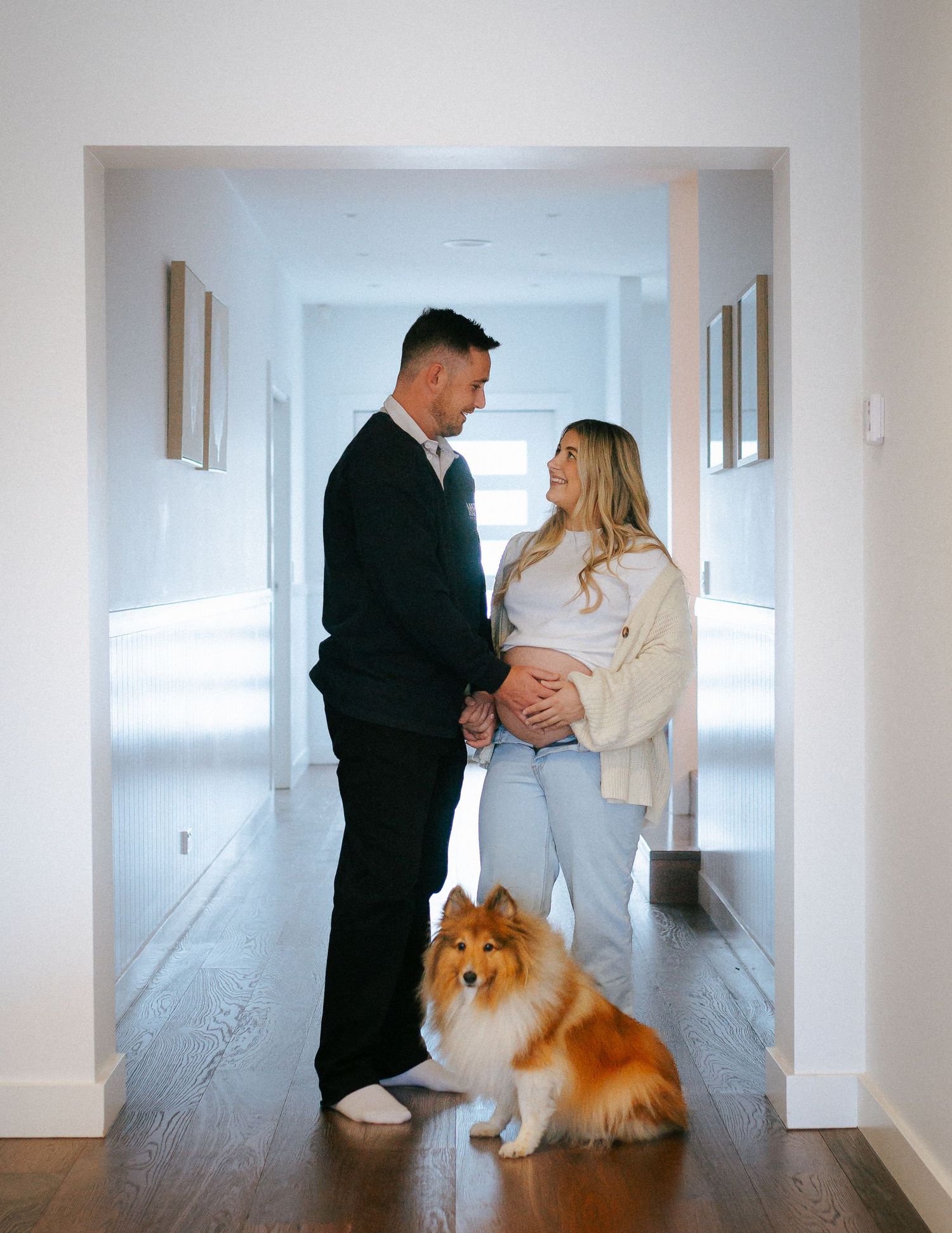 Pregnant couple holding baby bump in bright hallway with fluffy Collie dog sitting below.