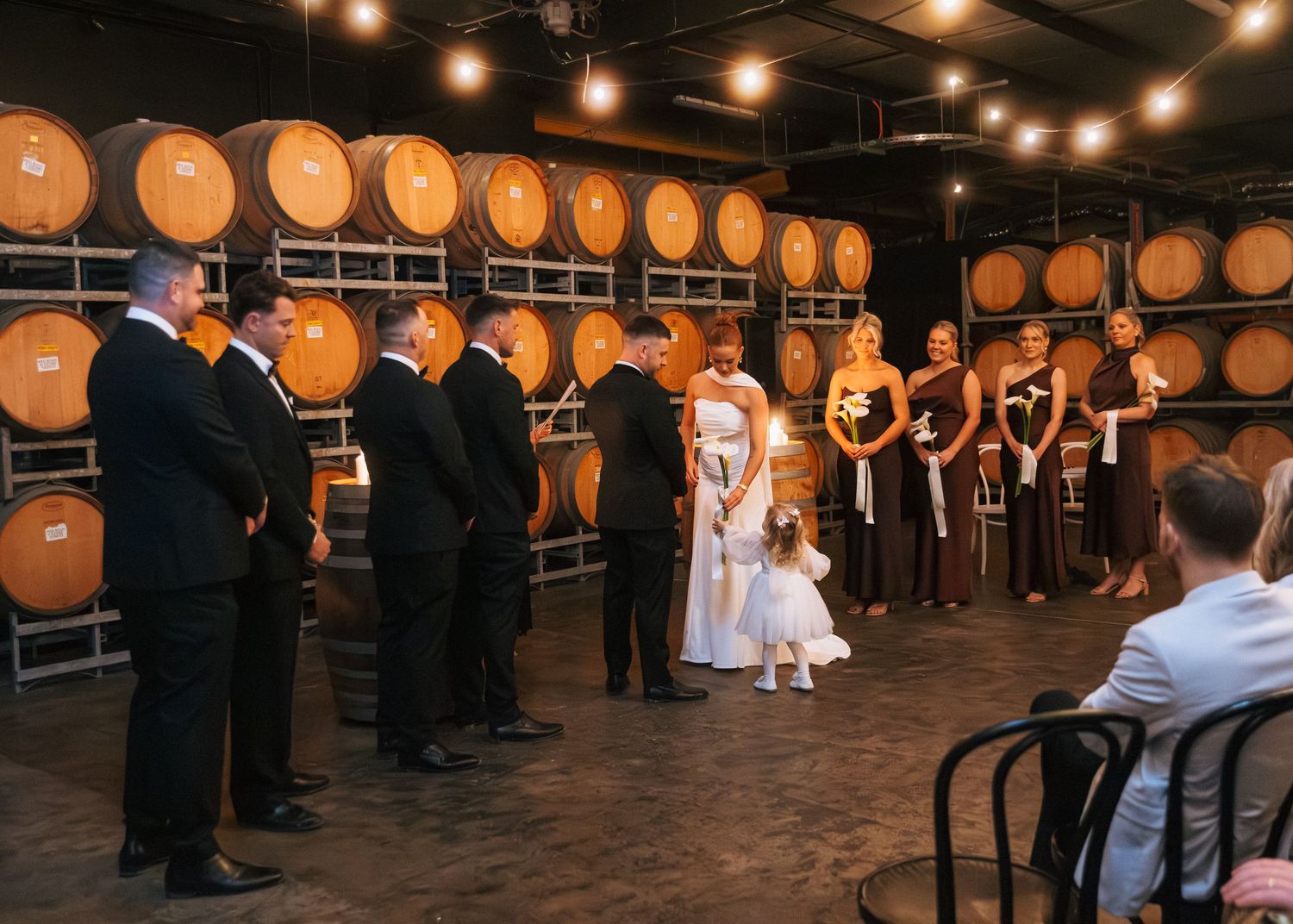 Wedding ceremony in a winery barrel room with bridal party standing among oak barrels under warm string lights.