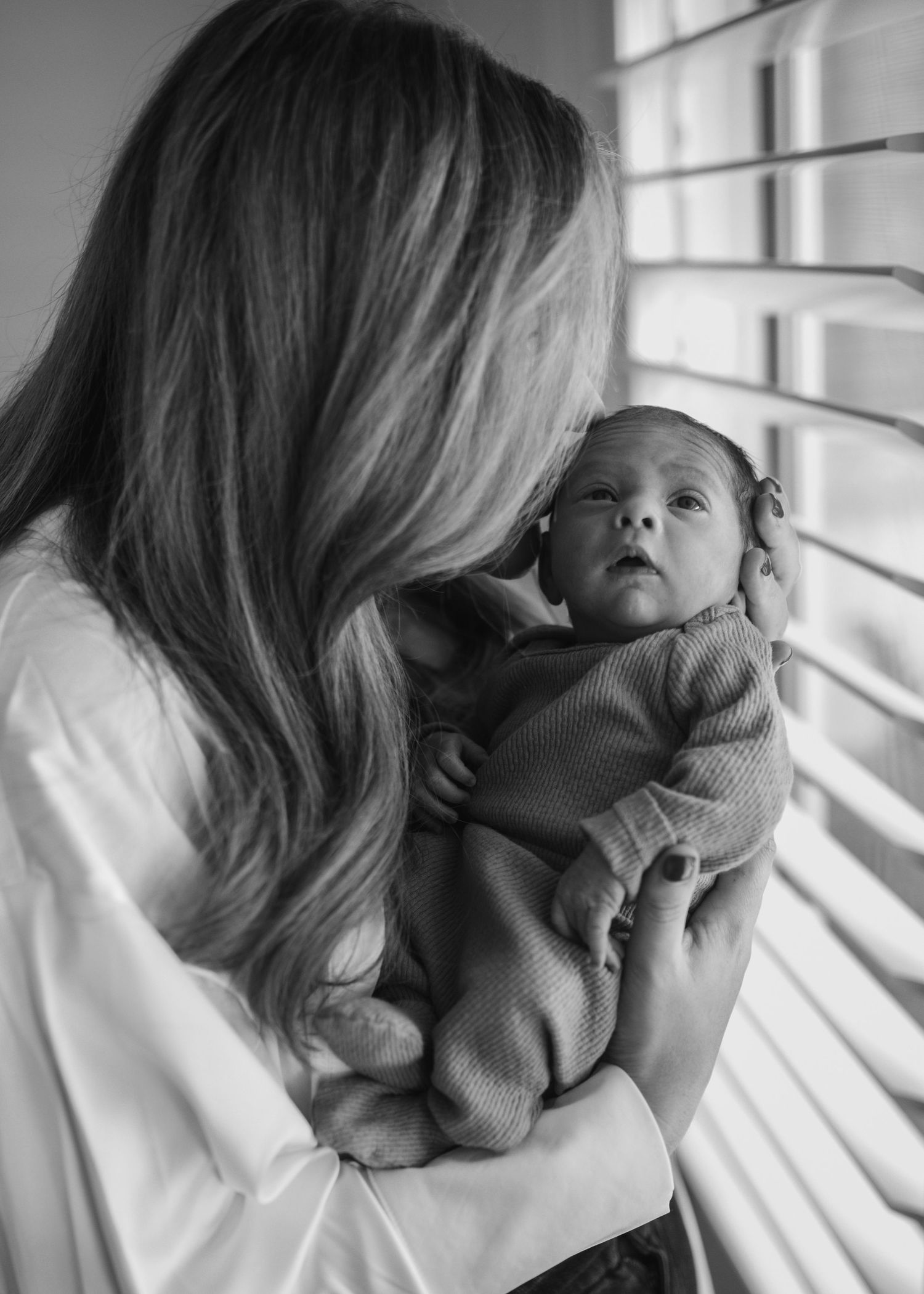 Mother with long blonde hair holding her newborn baby near a window in black and white.