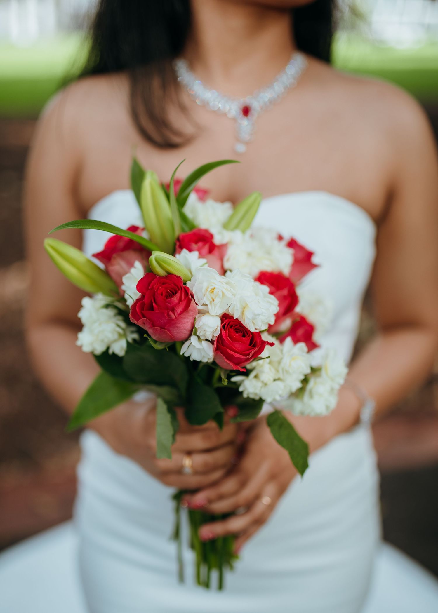 Close-up of bride holding elegant bouquet of red roses, white flowers, and lilies with diamond necklace visible.