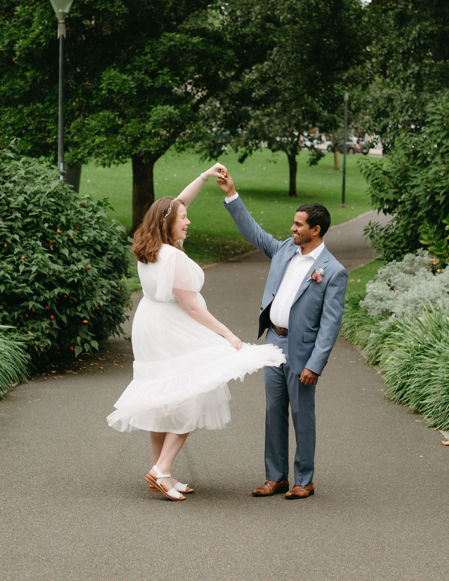 Groom twirls bride in a white tea-length dress on a park pathway during their joyful wedding photo session.