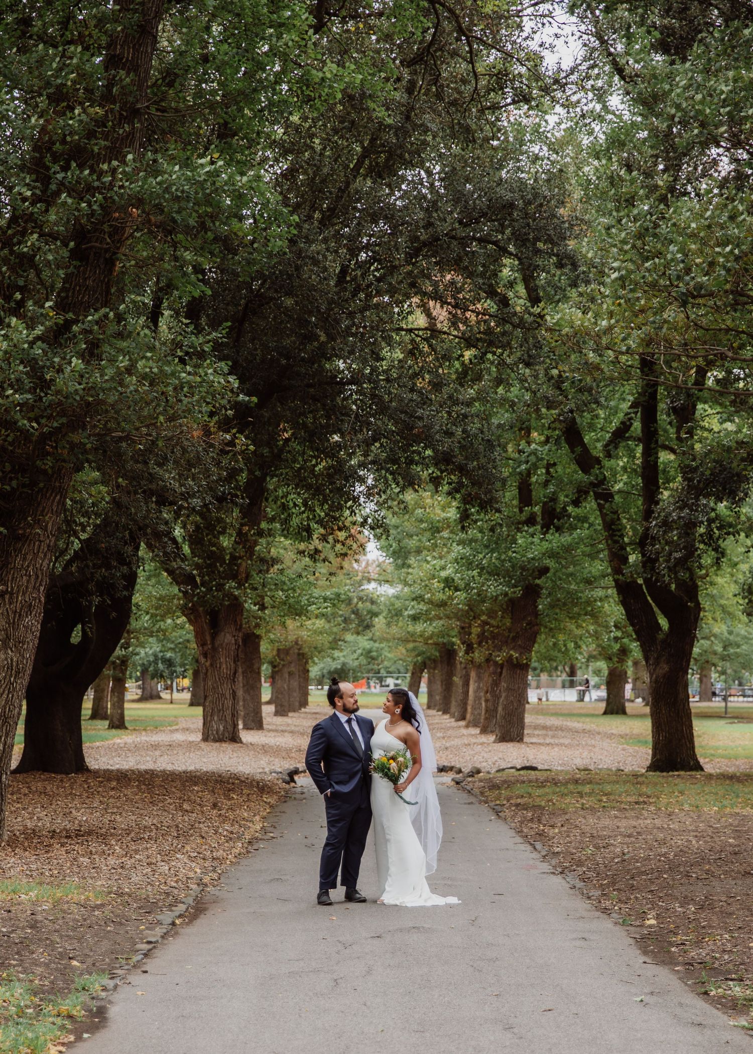 Bride and groom standing on a tree-lined park pathway in wedding attire.