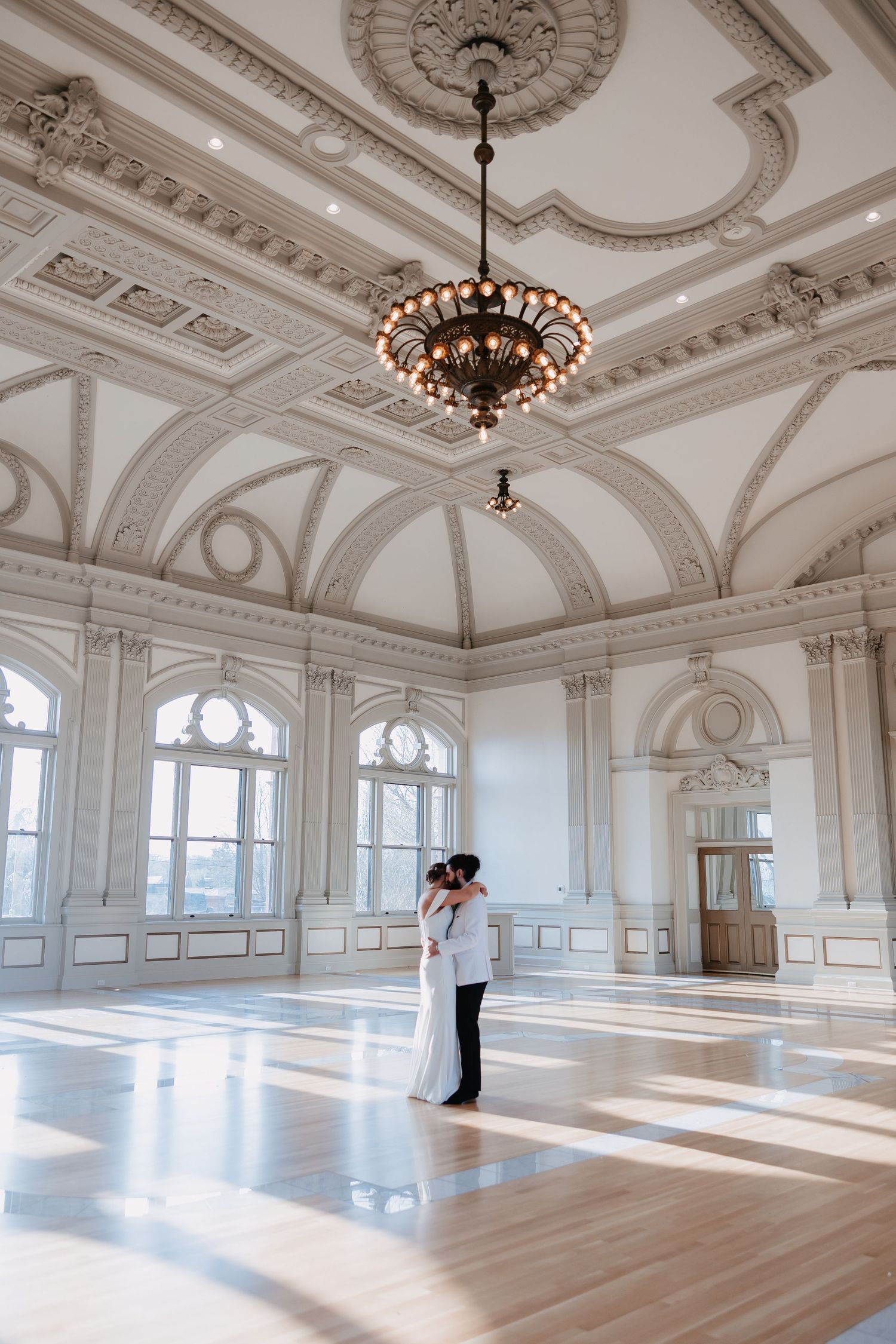 Bride and groom embracing in a stunning ornate ballroom with vaulted ceilings and chandelier. Steven's School Ballroom Wedding Photography