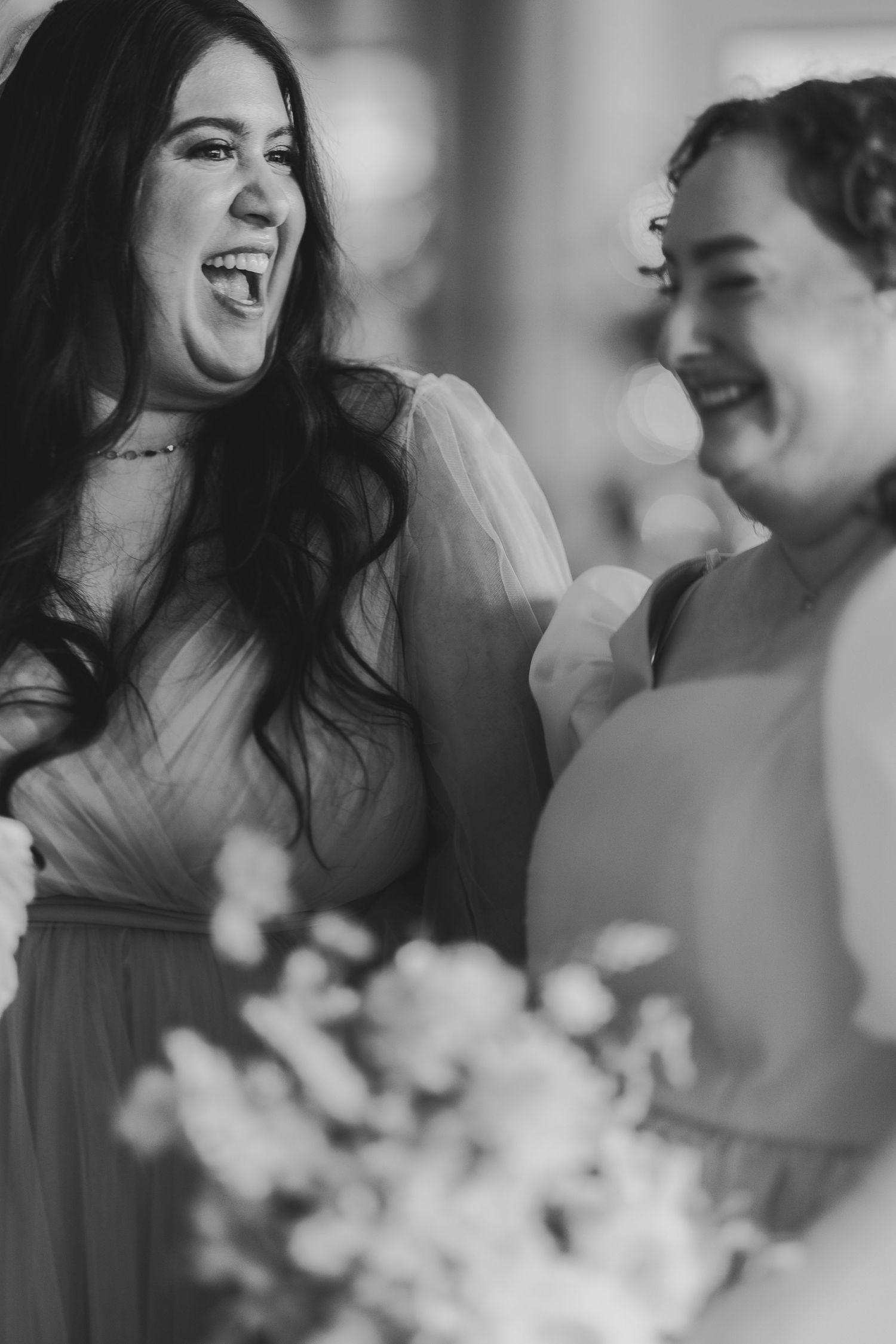 Black and white candid photo of two bridesmaids laughing joyfully together during a wedding celebration.