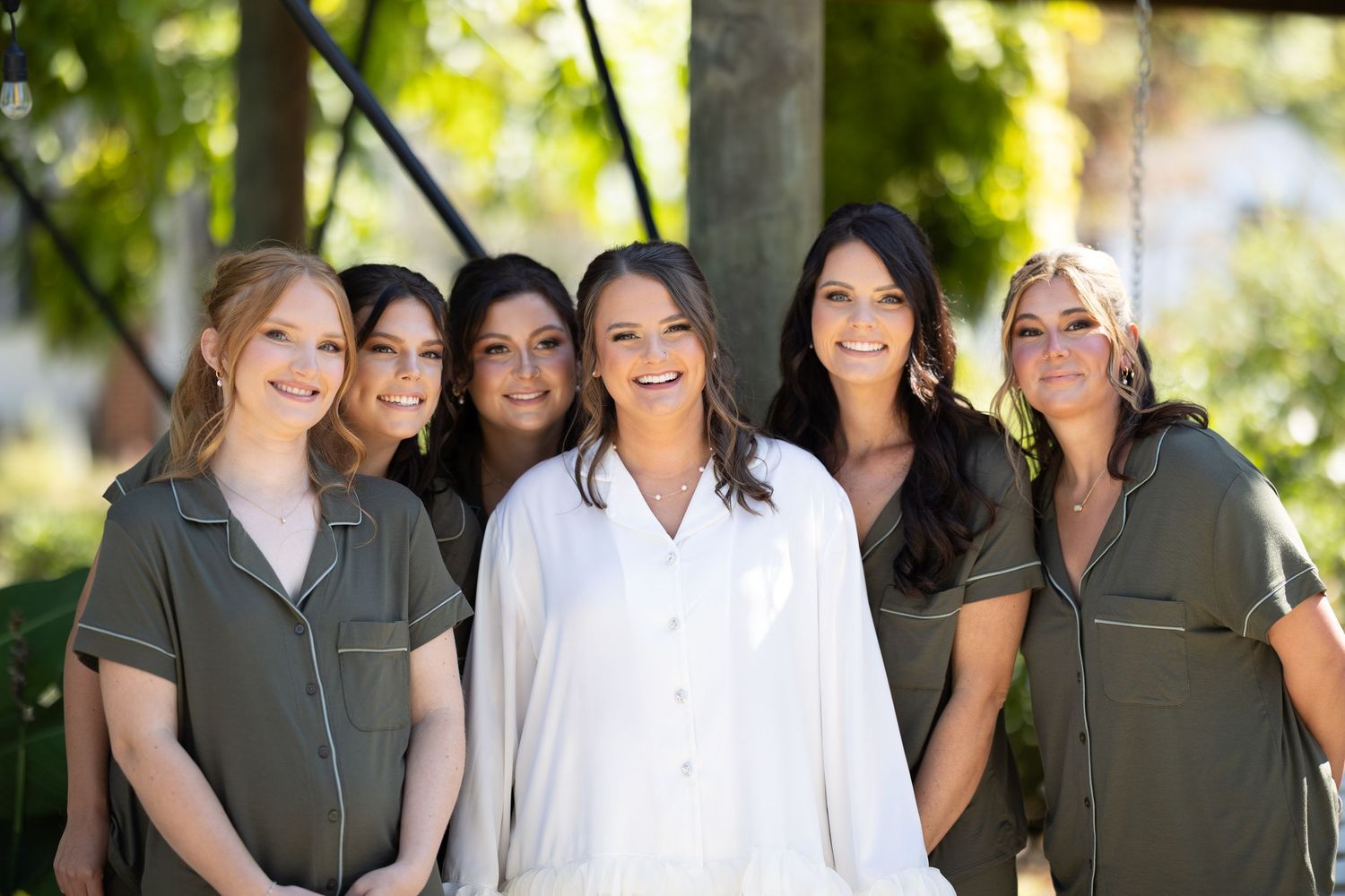 Bride in white robe smiles with five bridesmaids wearing olive green matching pajama sets outdoors.
