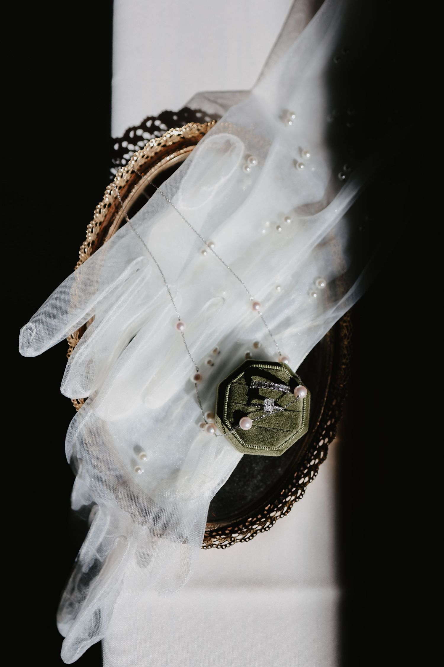 Elegant bridal flat lay featuring pearl-embellished gloves, delicate necklace, and vintage ring box on dark background.