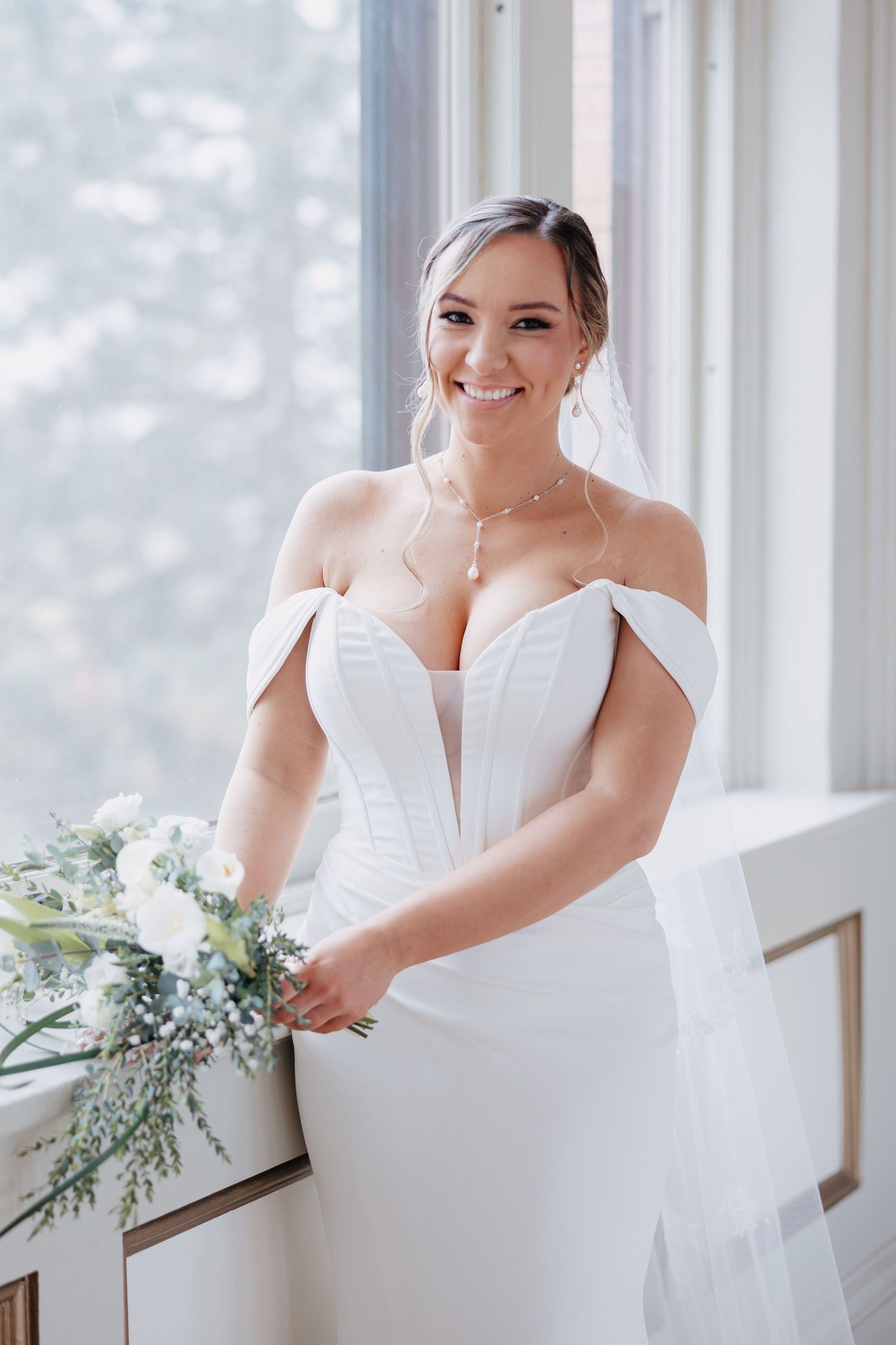 Radiant bride in off-shoulder corset wedding gown holding bouquet beside bright window at Steven's School Ballroom in Lancaster PA