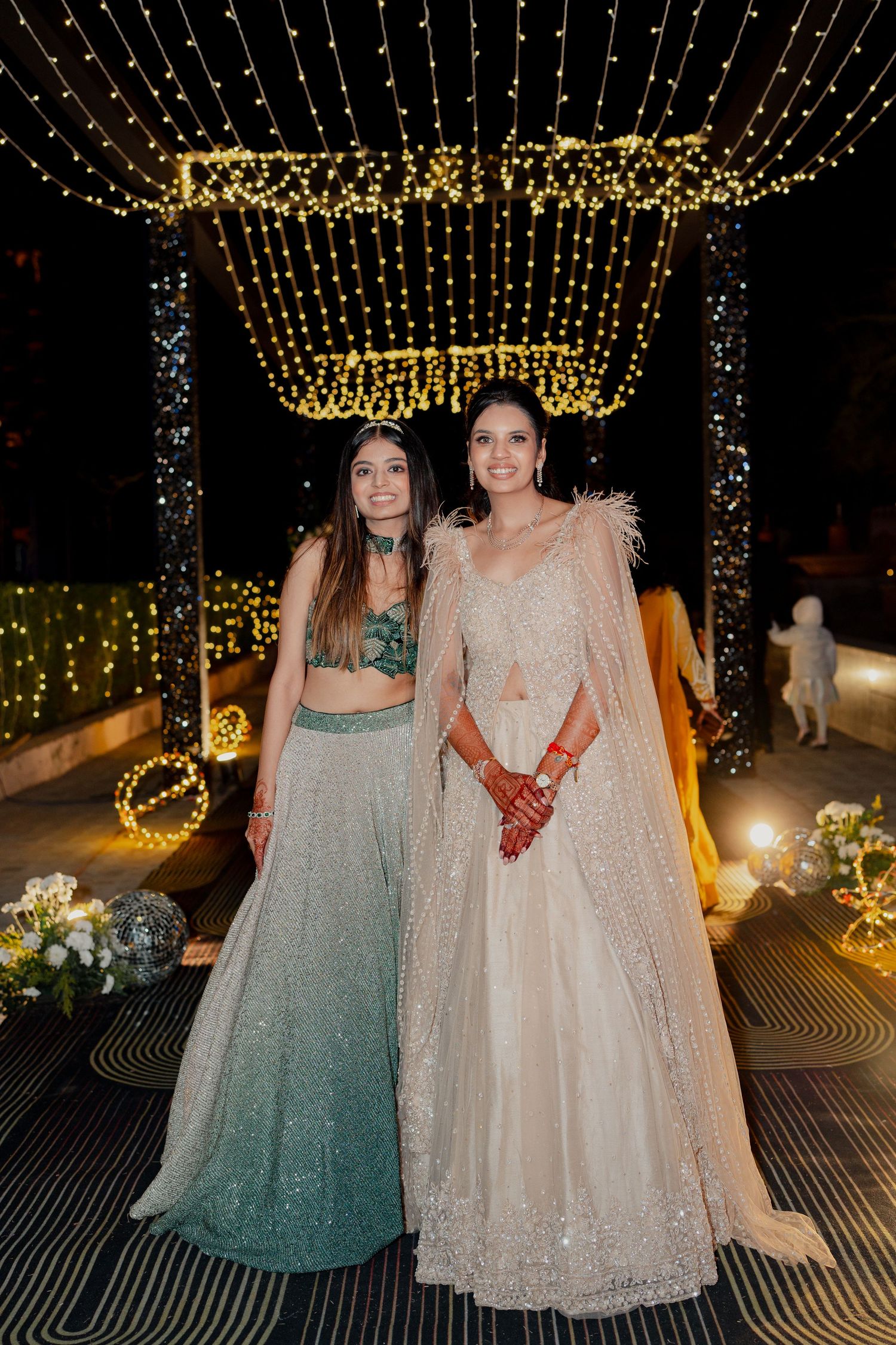 Two women in elegant Indian lehengas pose at a festive night event adorned with golden fairy lights.