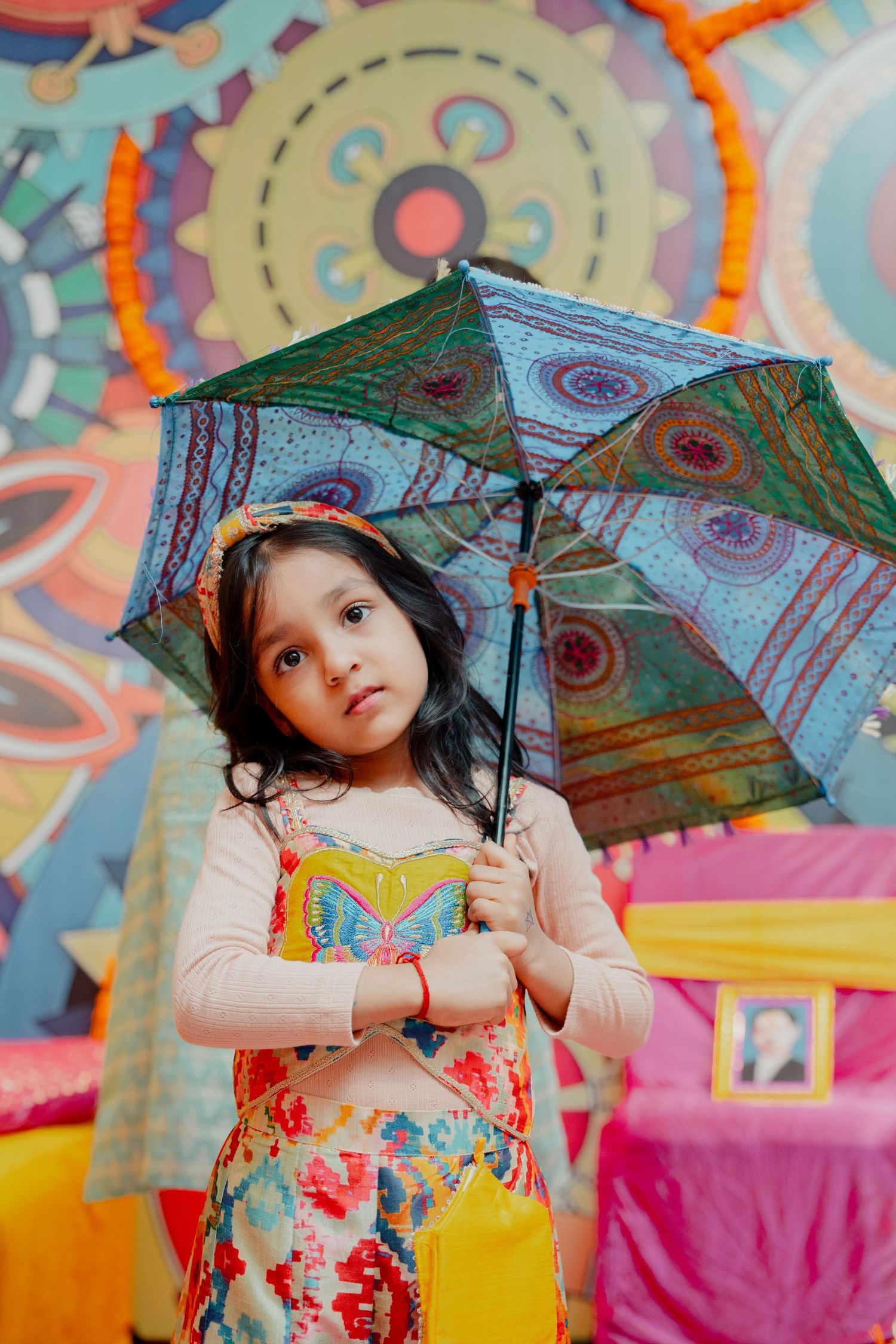 Young girl in colorful dress holding a decorative umbrella, standing before a vibrant painted backdrop in Eastern PA for an Hindu wedding