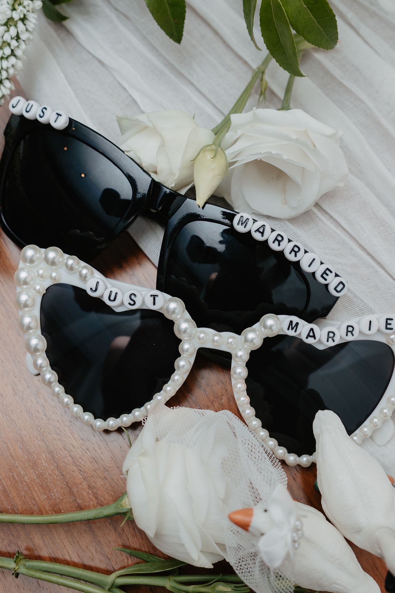 Two pairs of Just Married sunglasses decorated with pearls and beads surrounded by white roses on wood.