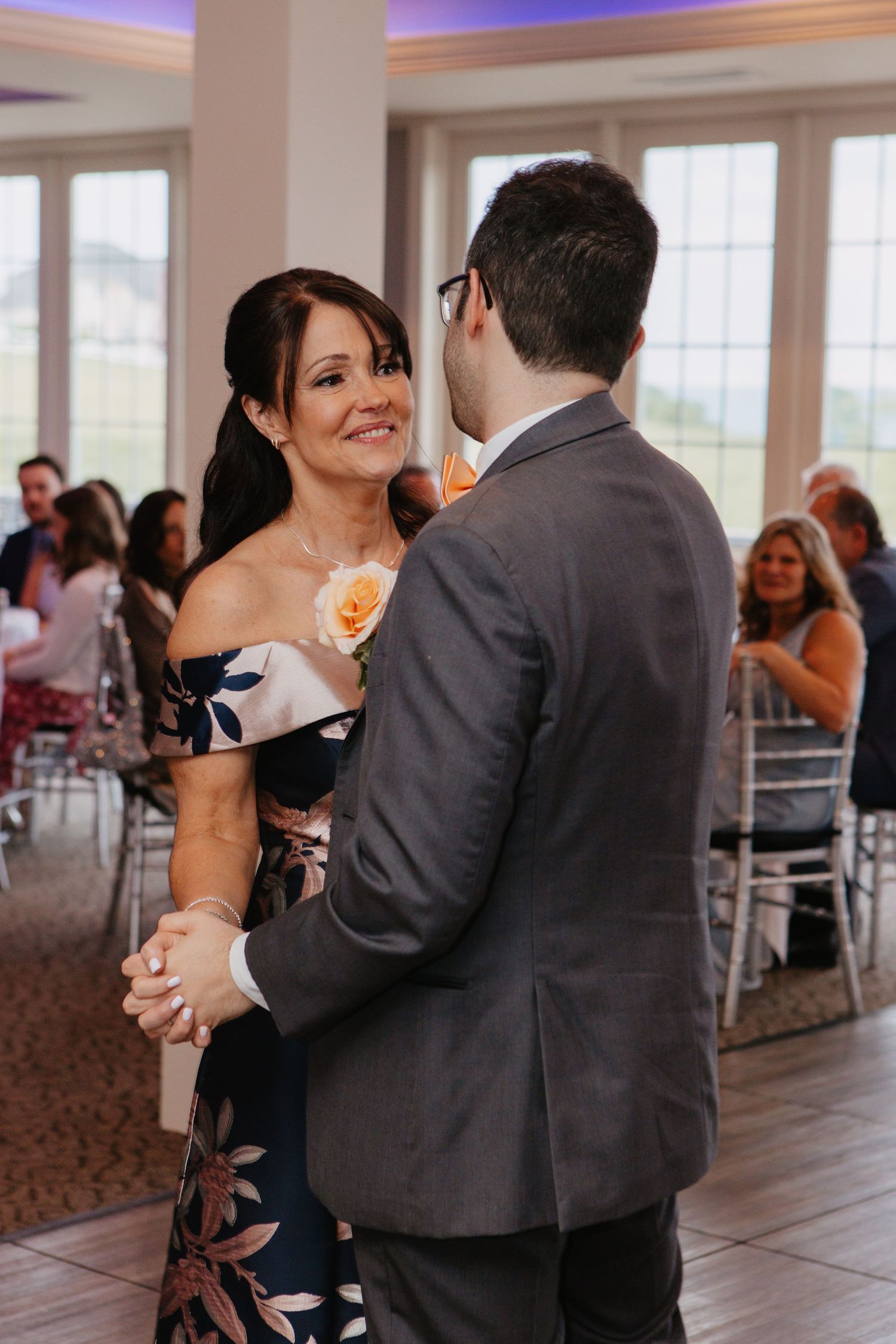 A woman in a floral dress dances with a man in a gray suit at a wedding reception, smiling warmly.