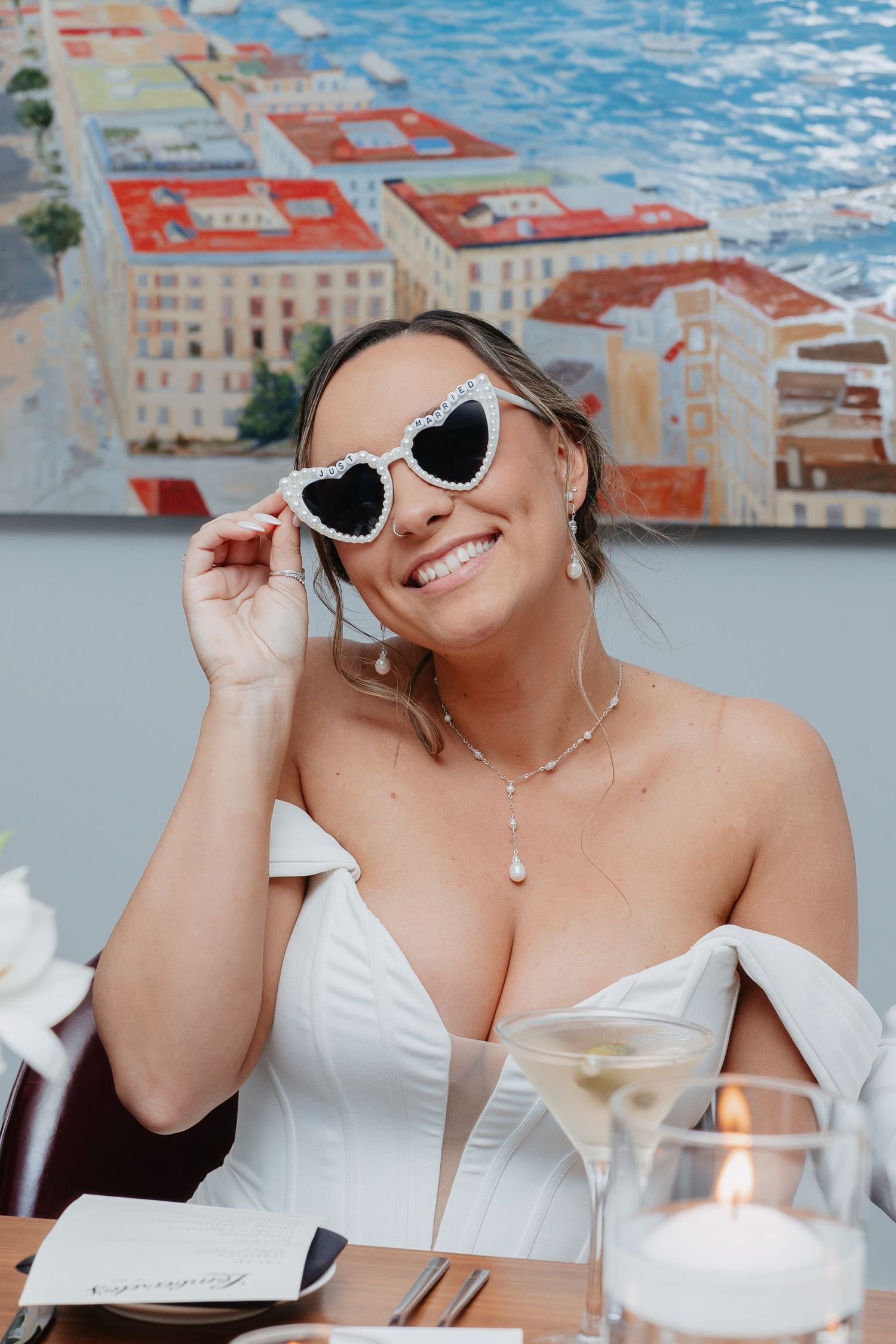 Smiling bride wearing heart-shaped pearl sunglasses in a white off-shoulder gown at her wedding reception.