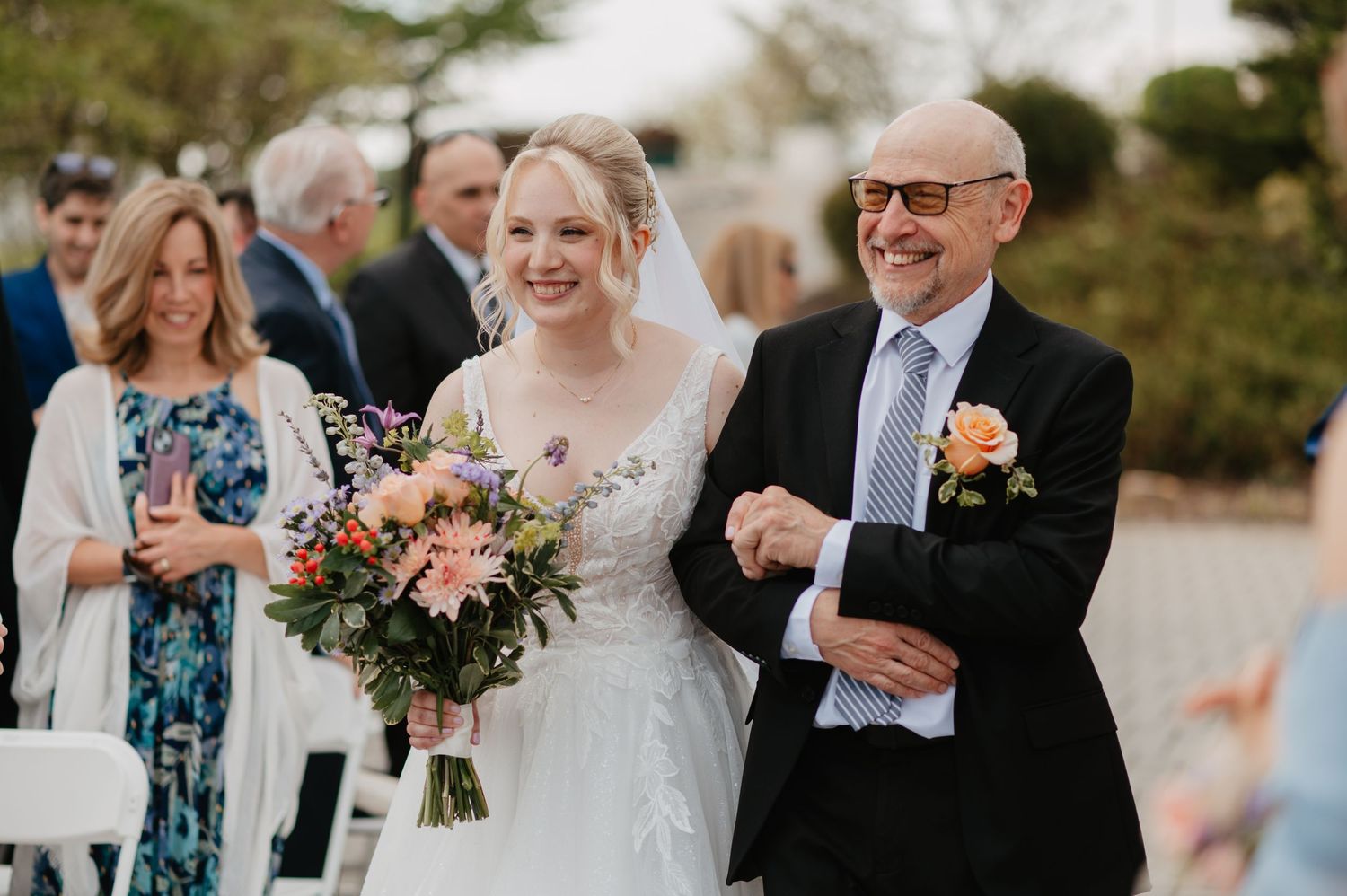 Bride in white gown holding colorful bouquet walks down aisle with bald man in suit at outdoor wedding ceremony.