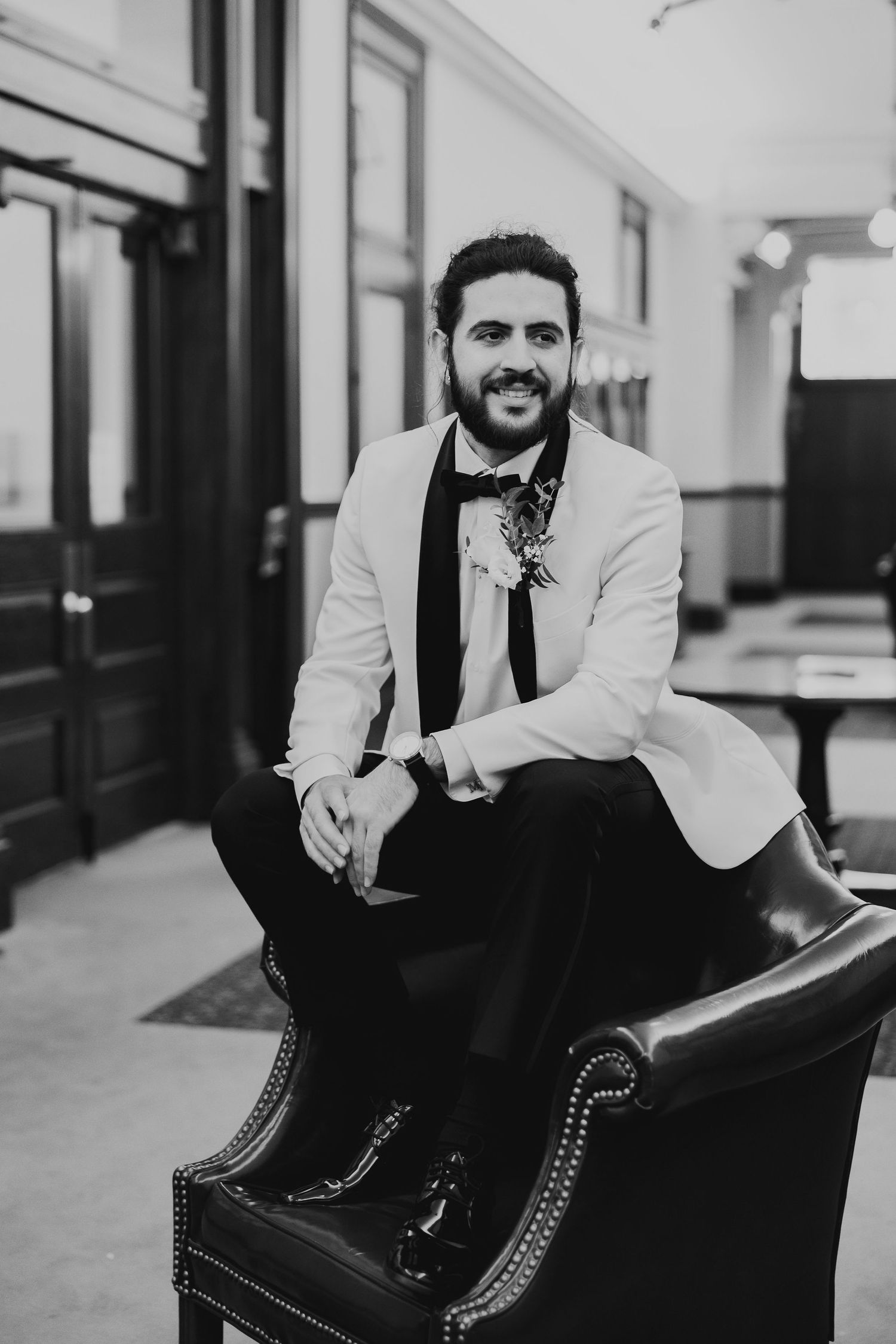 Dapper groom in white tuxedo jacket and bow tie seated in leather chair, black and white photo. Editorial Groom Coverage