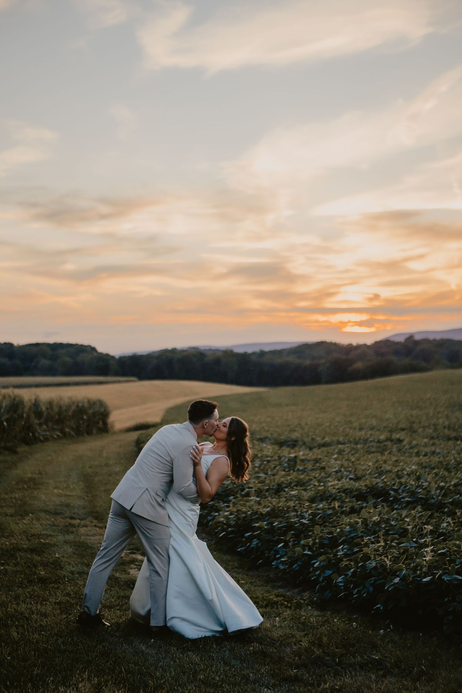 Bride and groom share a romantic dip kiss in a lush green field during a stunning golden sunset.