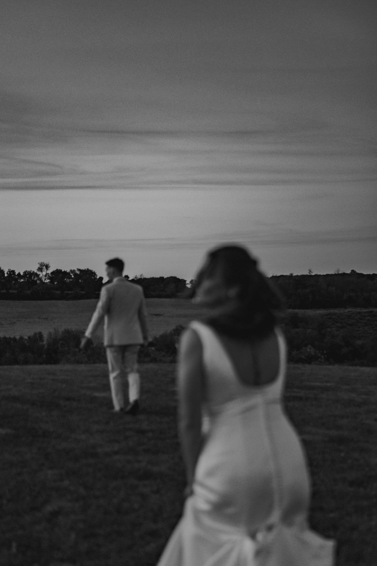 Artistic black and white film photo of bride and groom walking across grassy field at twilight in Bucks County PA.