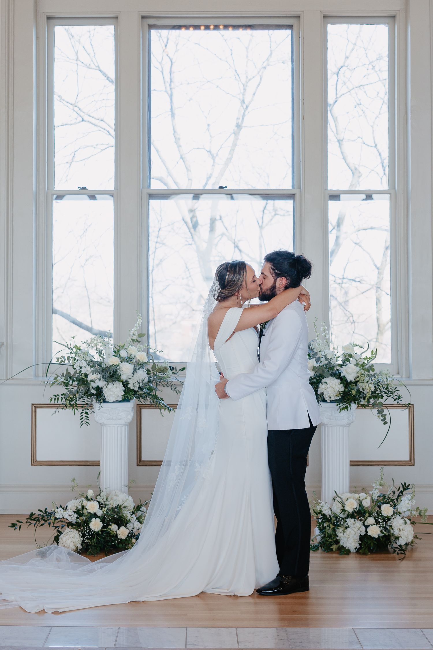 Bride and groom sharing first kiss at altar surrounded by white floral arrangements and large windows.