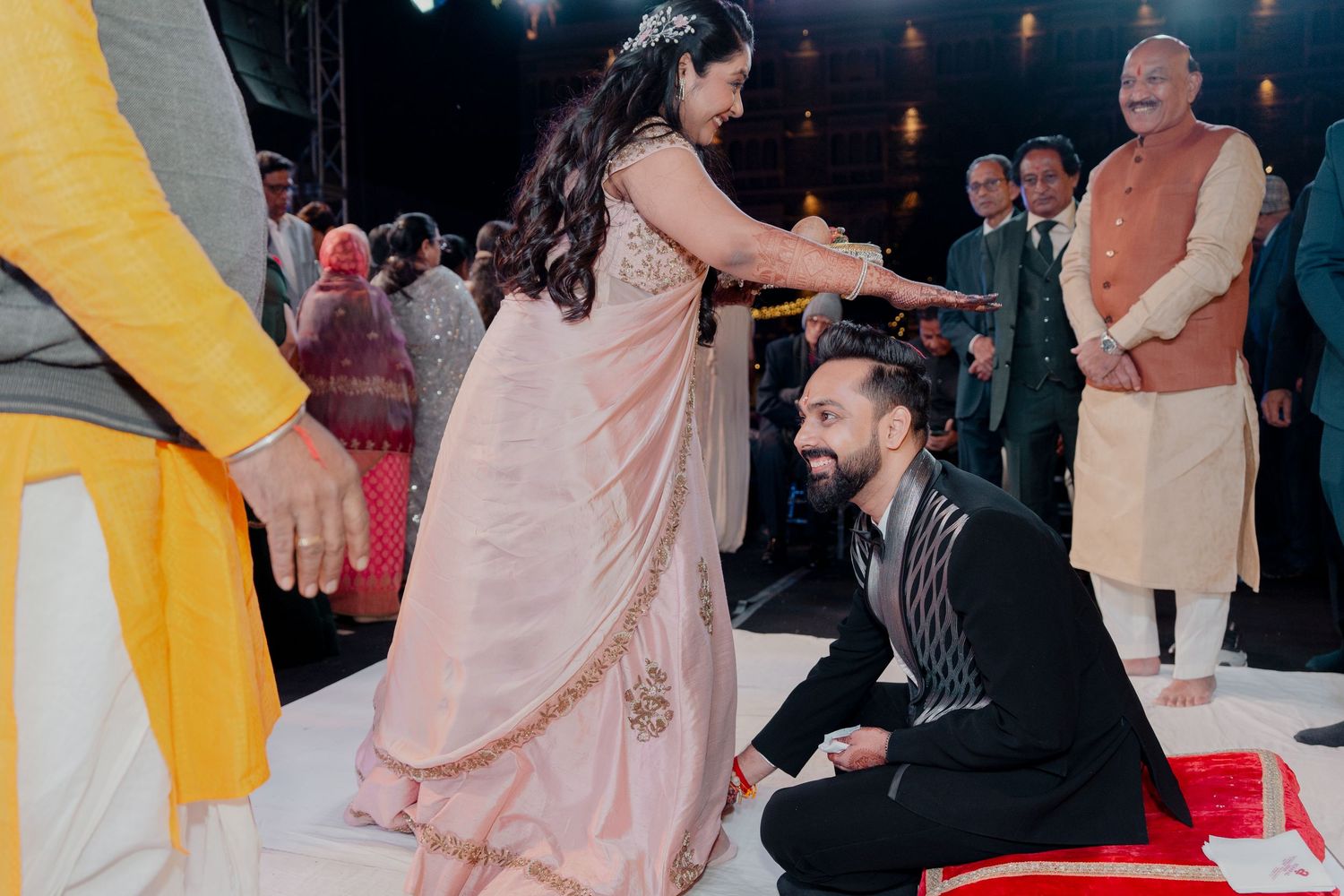 Man kneeling before woman in pink saree at elegant Indian wedding ceremony with guests in background.