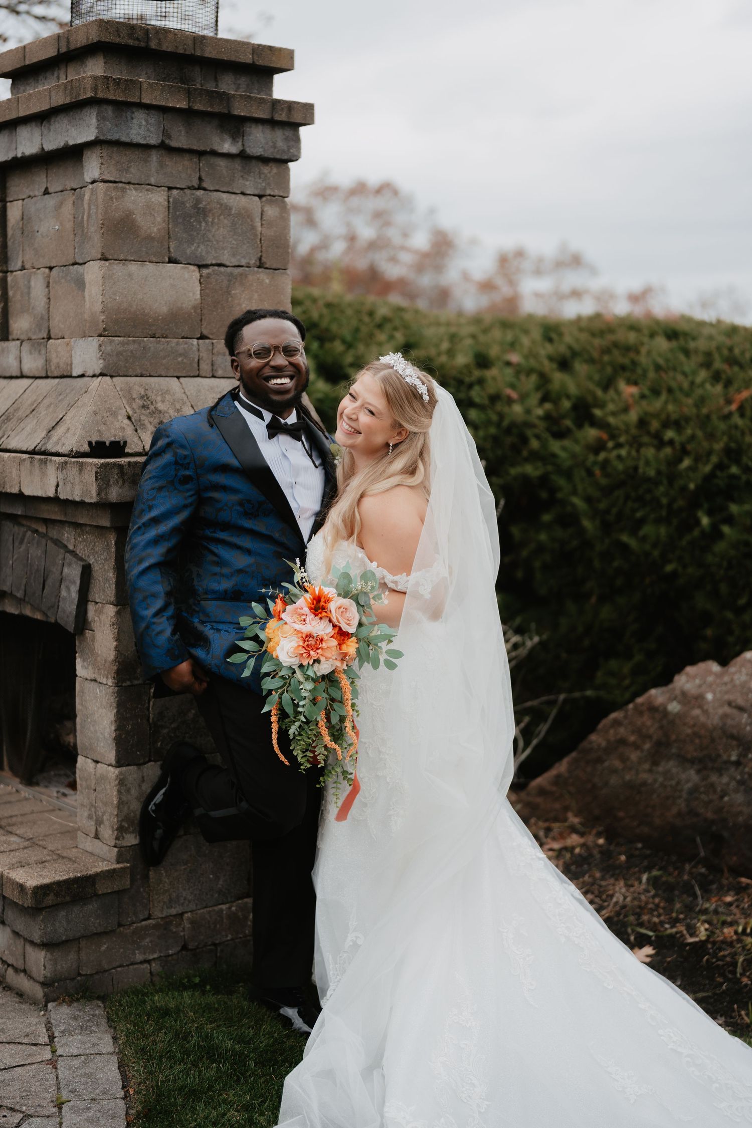 Smiling newlywed couple posing by stone outdoor fireplace, bride in white gown with orange bouquet, groom in navy tuxedo. Film and Digital