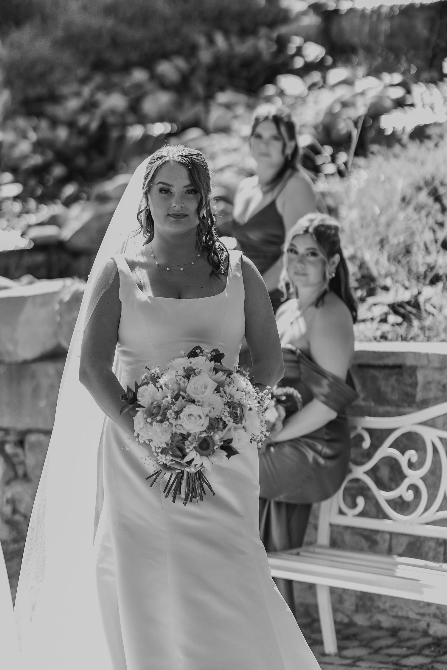 Elegant bride in a white gown holds a floral bouquet with bridesmaids in a black and white wedding photo.