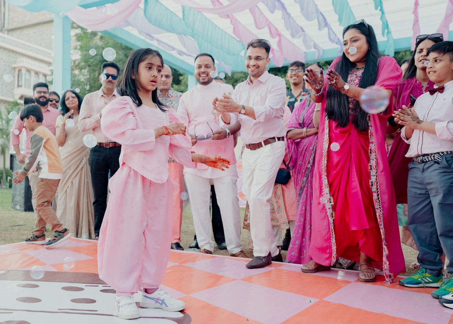 Young girl in pink outfit dancing at outdoor wedding celebration while guests in colorful attire watch and clap.
