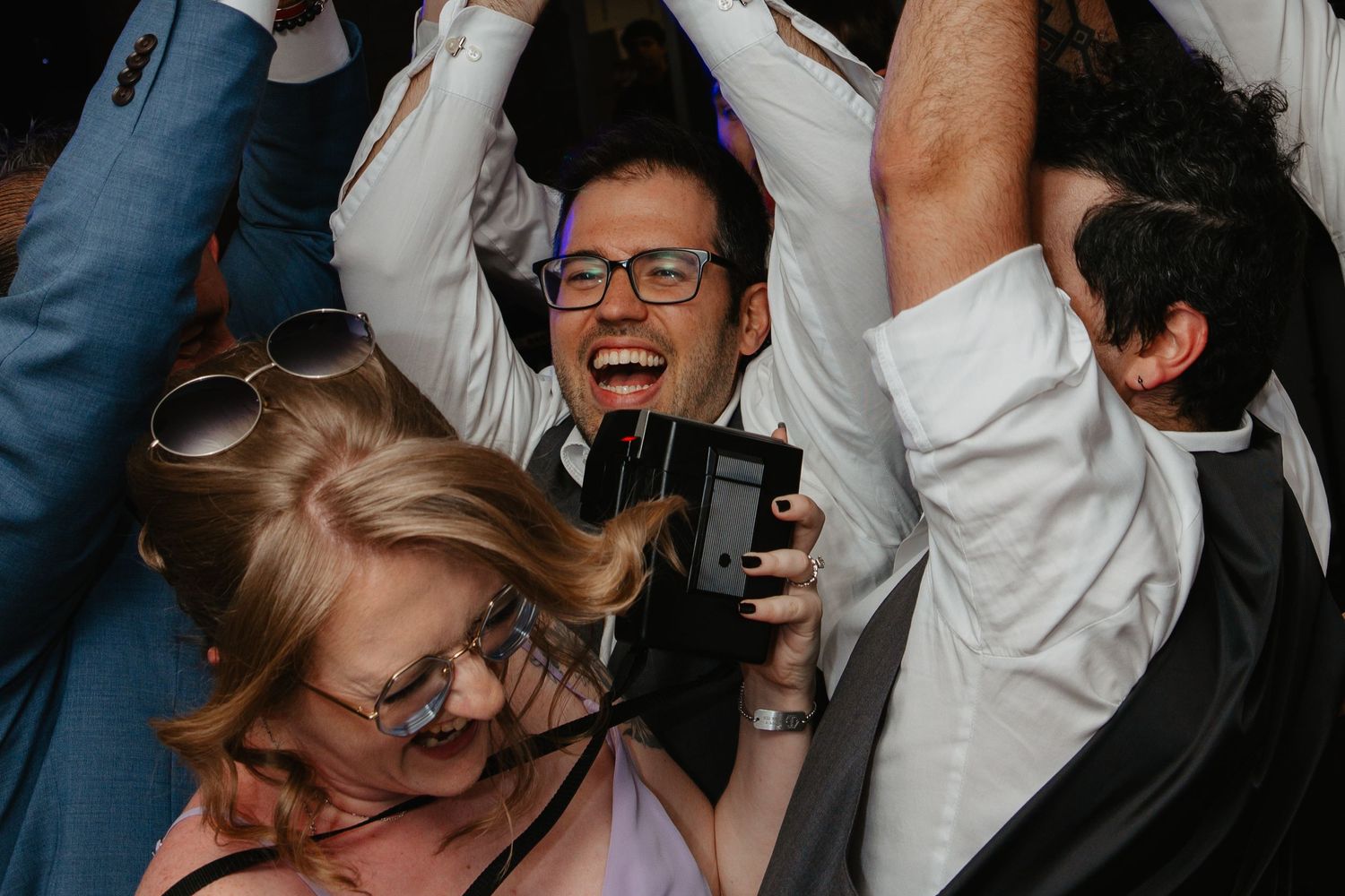 Joyful wedding guests dancing and celebrating, man holding camera smiling at lively reception party.