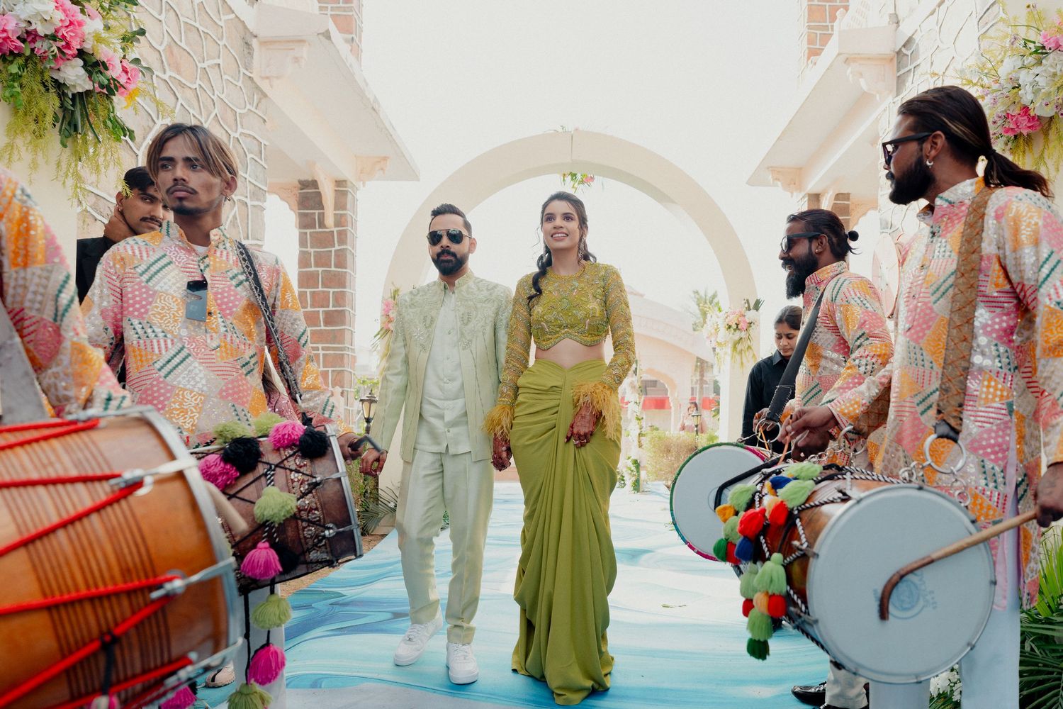 Couple walks through floral arch with dhol players in colorful outfits at vibrant Indian wedding celebration.