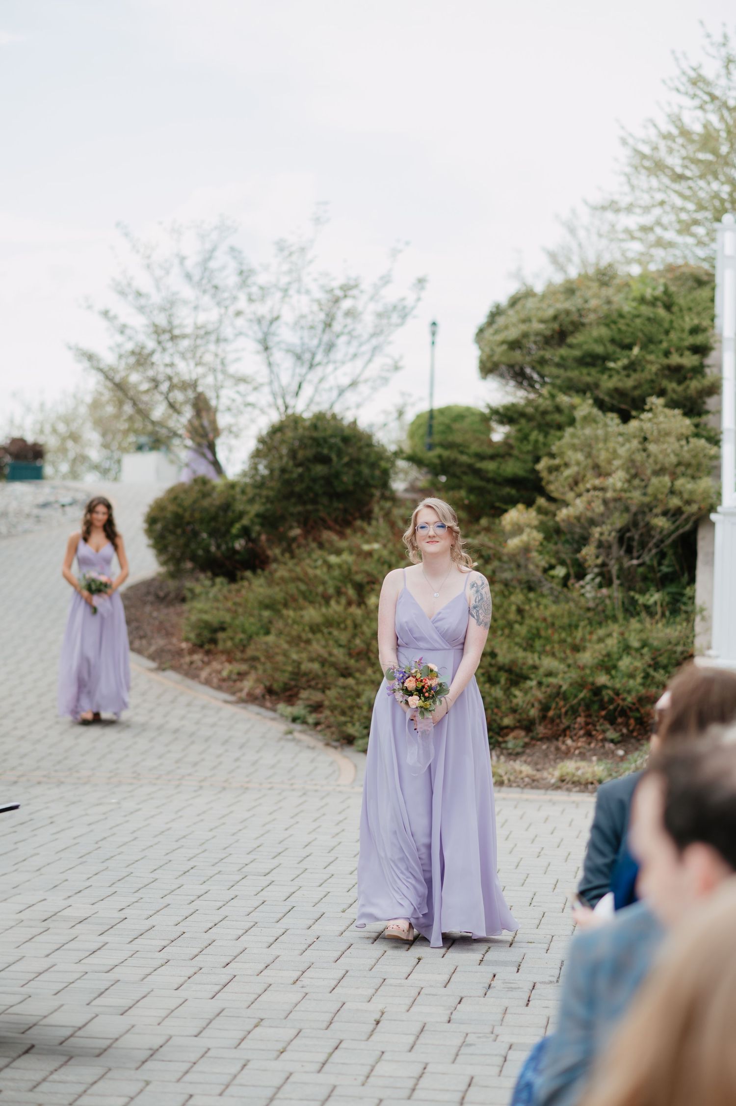 Bridesmaids in lavender gowns walk down an outdoor cobblestone aisle carrying colorful bouquets at a garden wedding.
