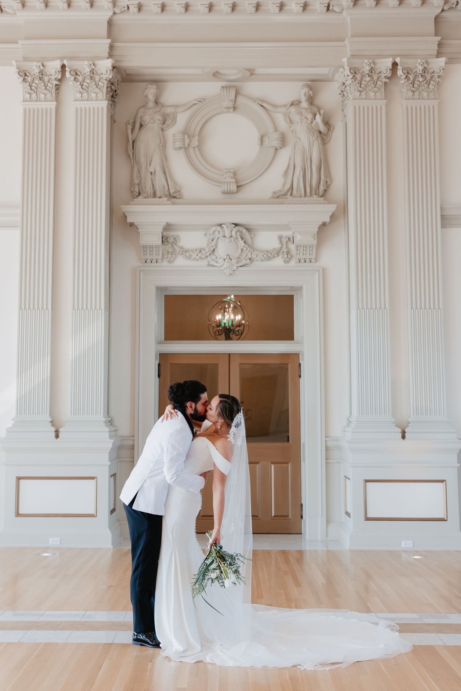 Bride and groom share a romantic kiss in front of ornate white columns and classical architecture at their wedding venue.